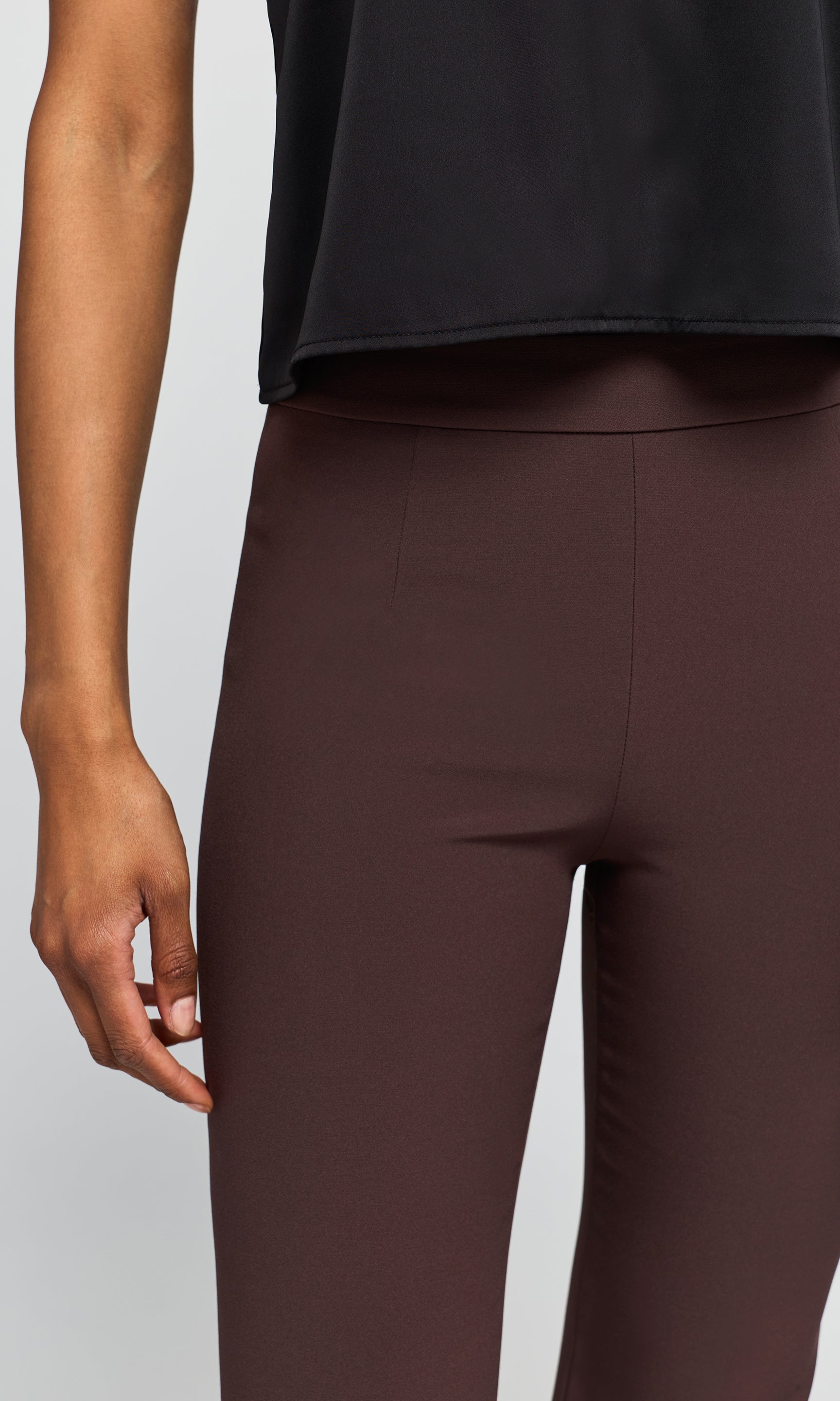 Close-up of a model wearing dark brown leggings and a black sleeveless top against a light gray studio background.