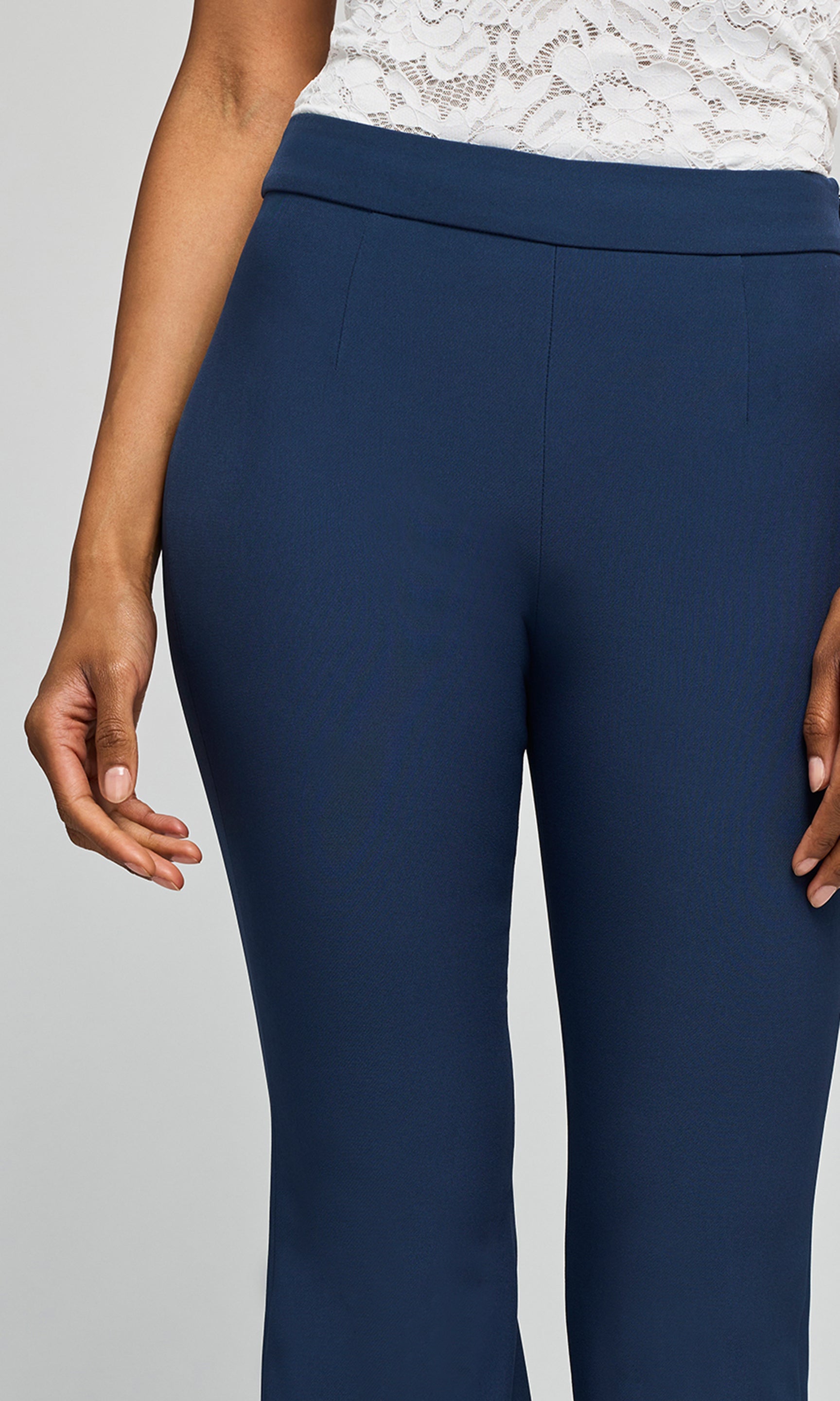 A close-up of a model wearing navy blue pants with a white lace top, on a plain background.