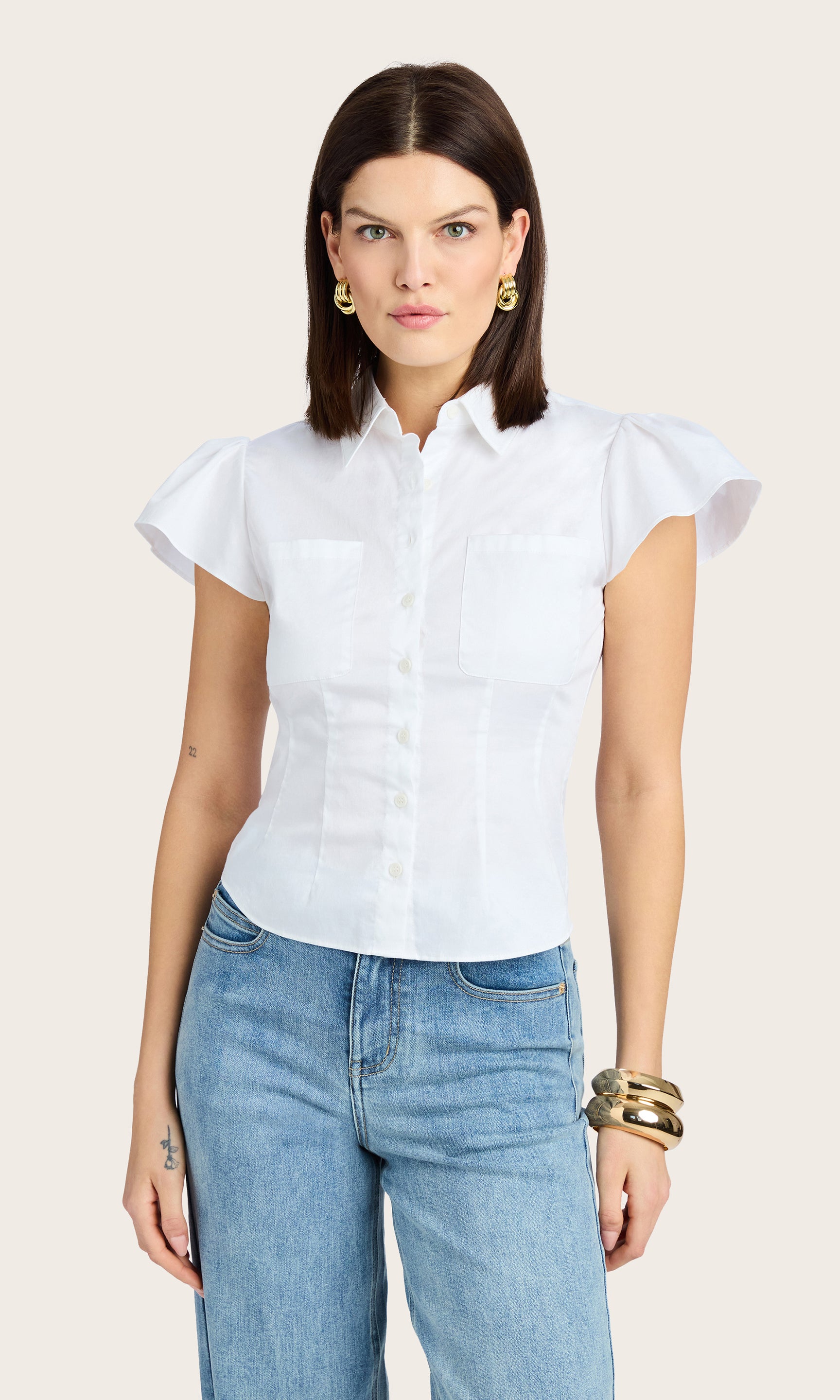 front image of model wearing the francesca poplin button down shirt with short flutter sleeves paired with blue jeans, accessorized with a gold bracelet and earrings