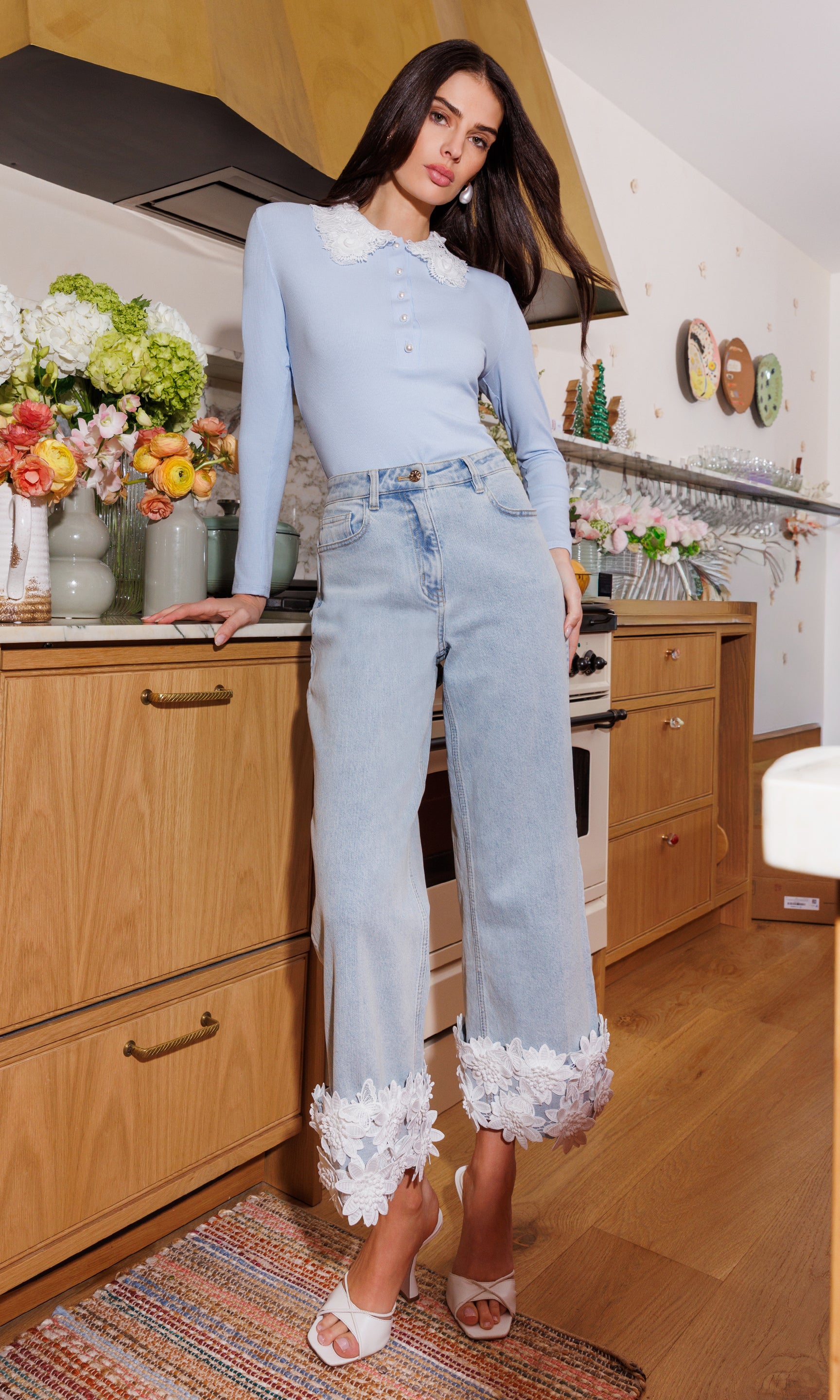 In a full body front view, the model wears the Paloma Lace Combo Jeans in light wash denim, showcasing the dramatic lace cuff detail in a styled kitchen setting.
