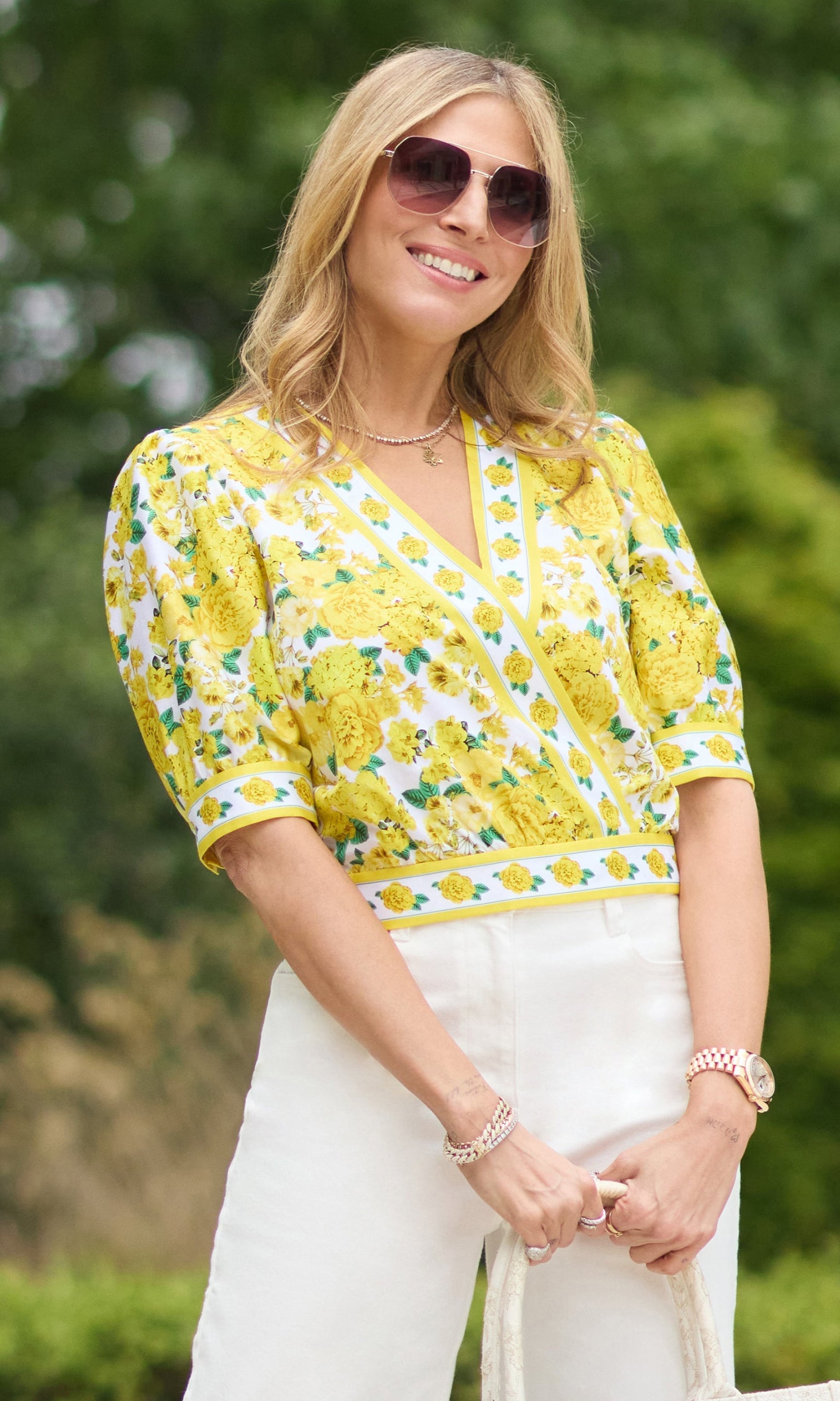 image of founder Roni posing in a garden wearing the wearing the Rema Floral top in a white/yellow pattern paired with white jeans