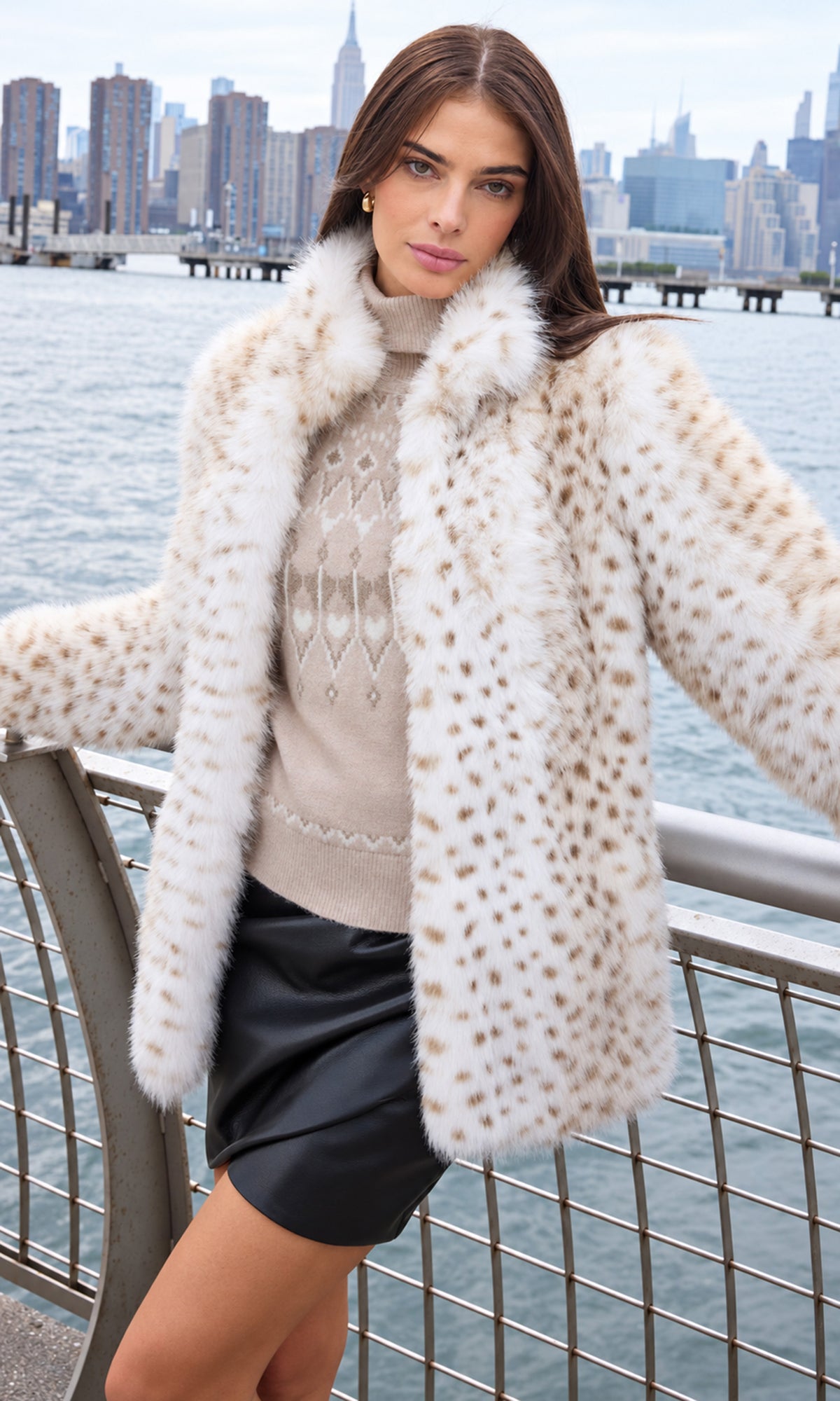 A woman models a cream faux fur coat with brown spots, worn over a beige knit sweater and a black leather mini skirt, with a city skyline and water in the background.