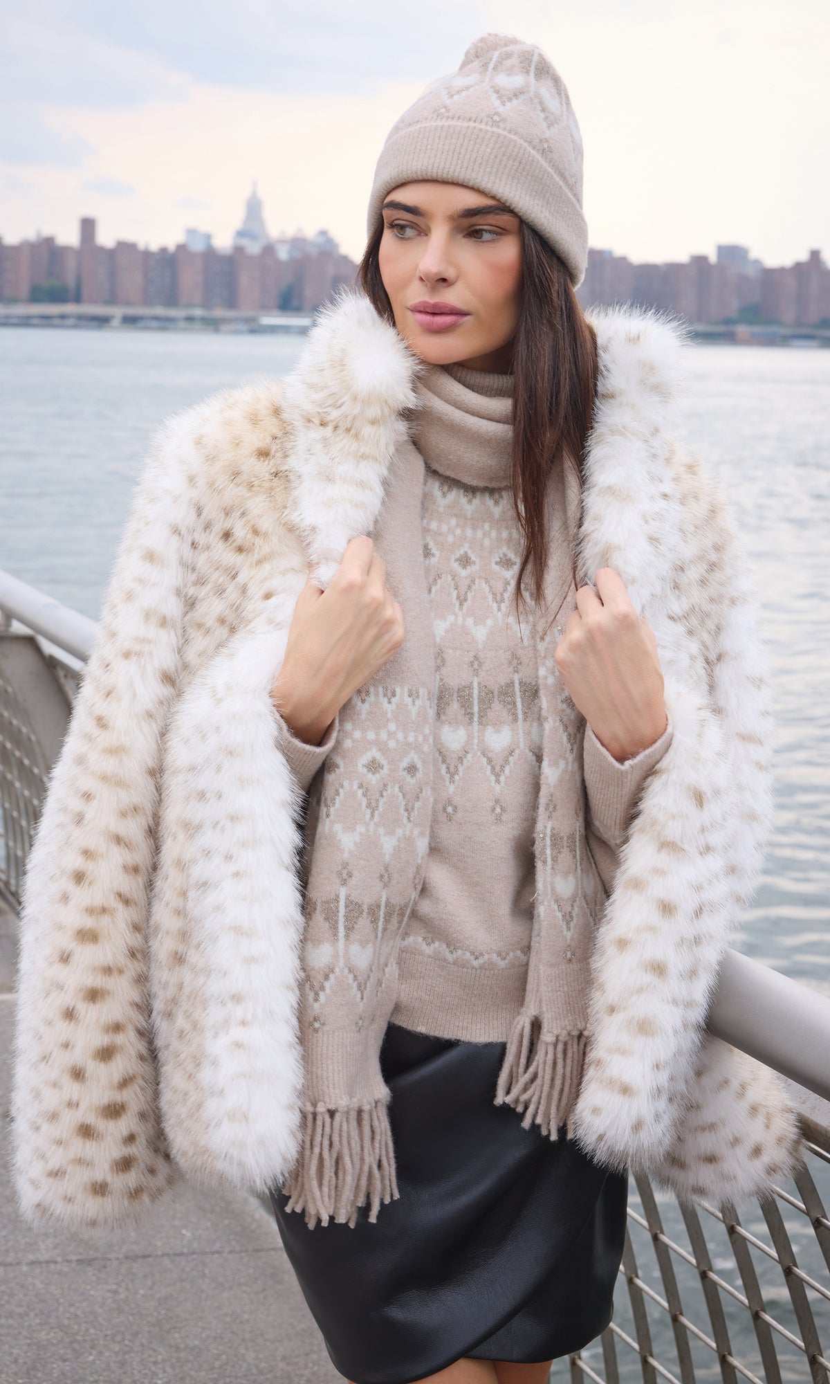 A woman wearing a beige knit beanie, a cream and brown spotted faux fur coat, a beige knit sweater, and a matching scarf, standing outdoors with a city skyline and water in the background.