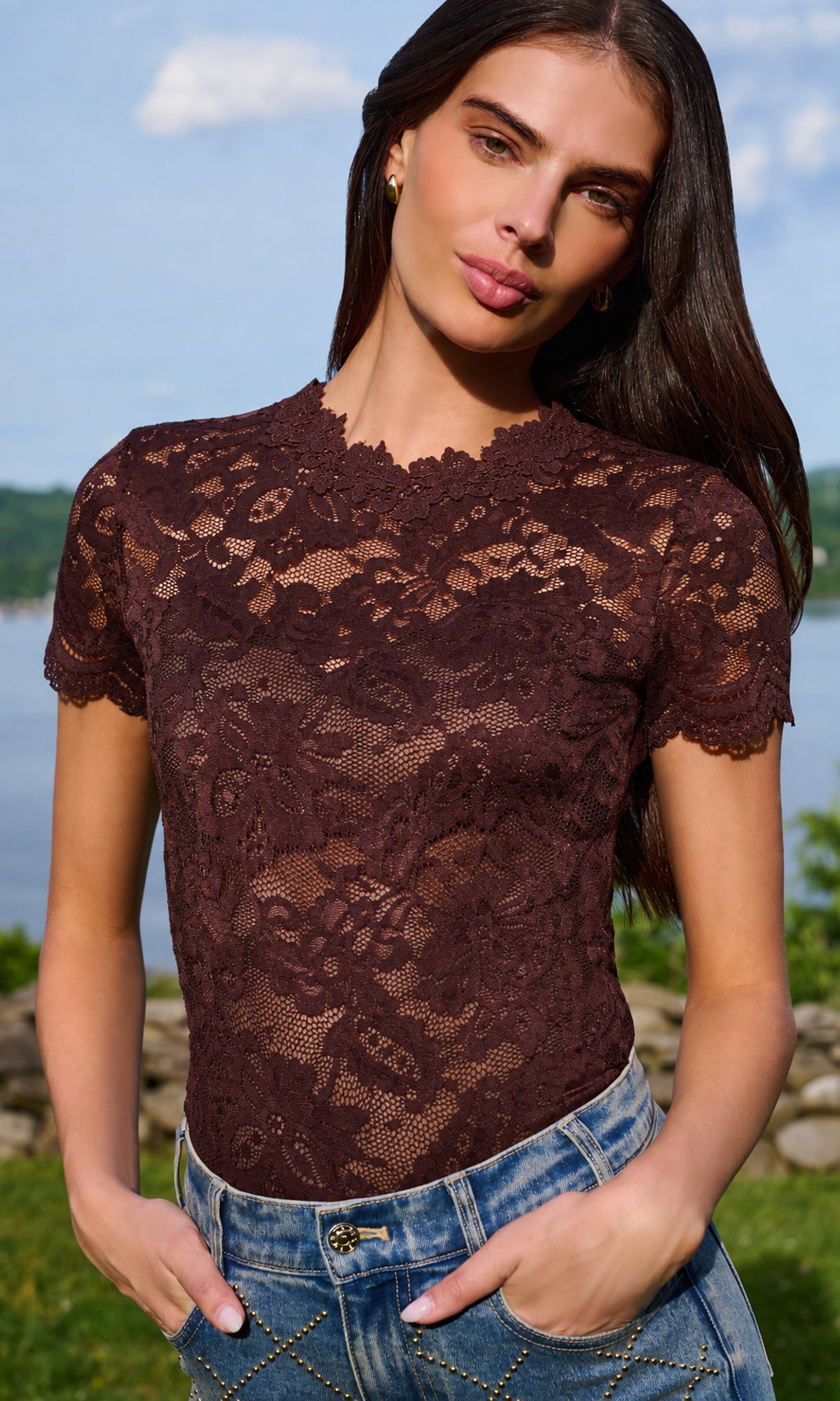 Model wearing a short-sleeve lace top in dark brown, paired with blue jeans, standing outdoors.