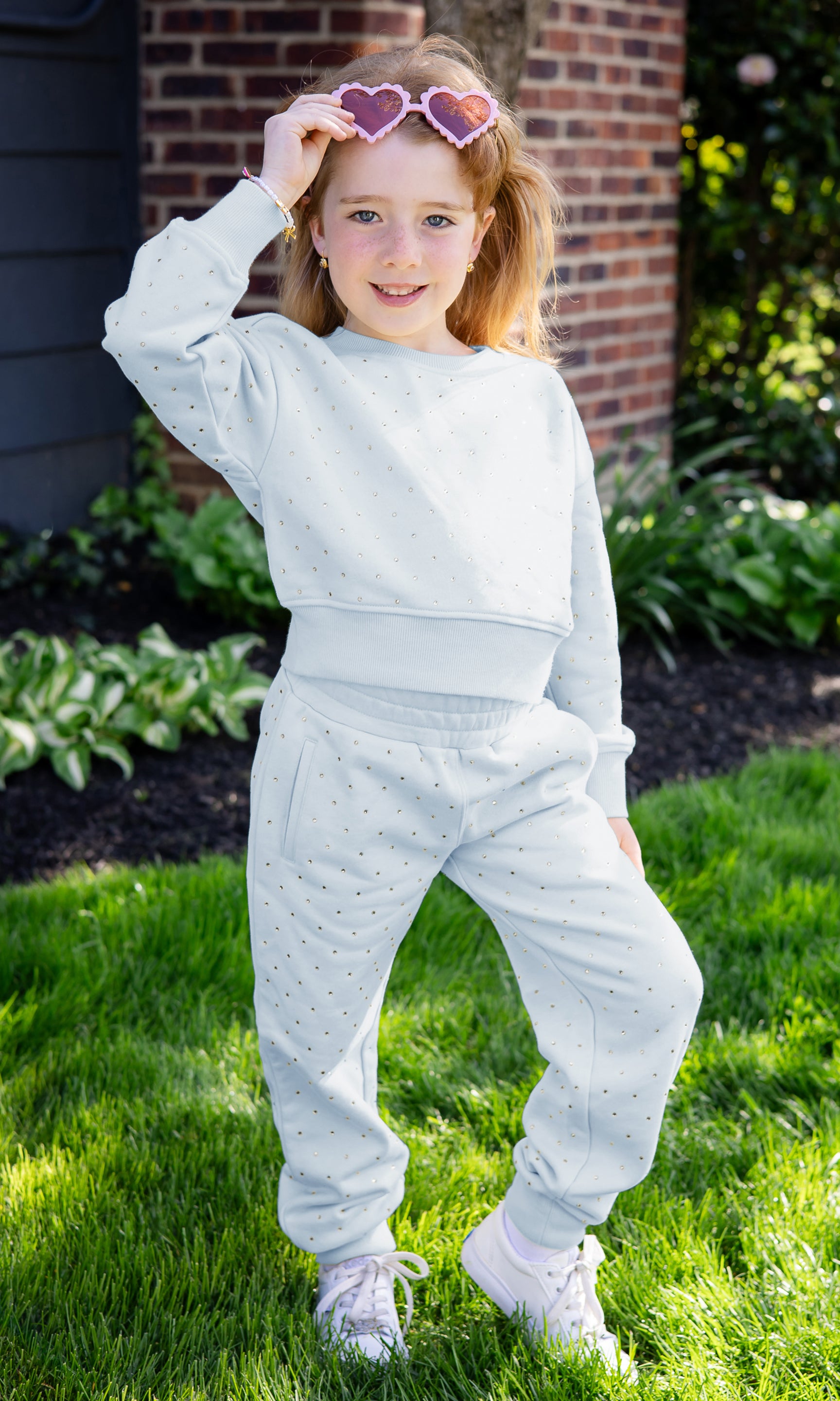 Child wearing a light blue outfit outdoors with a brick wall and greenery in the background