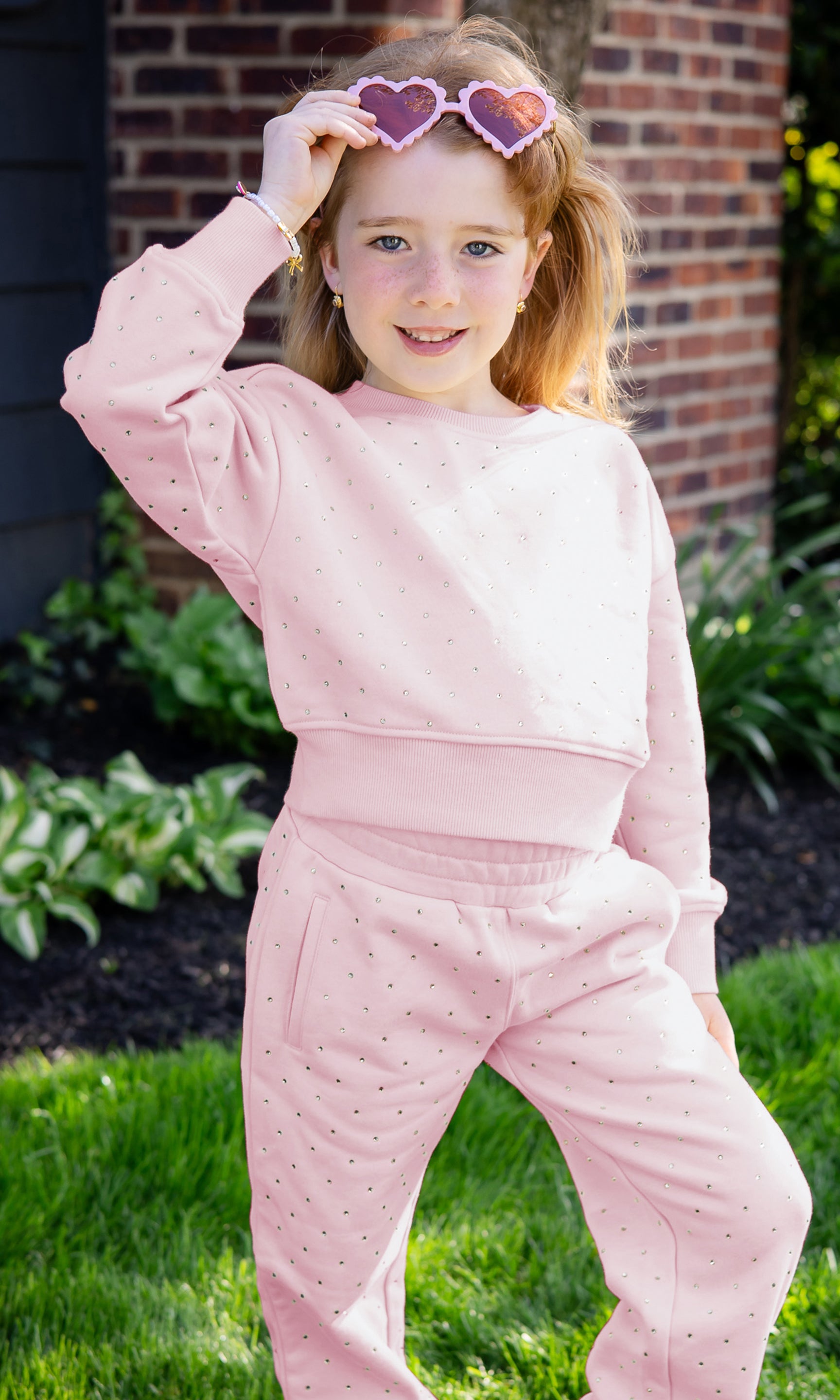 Young girl wearing a pink outfit with heart-shaped sunglasses outdoors.