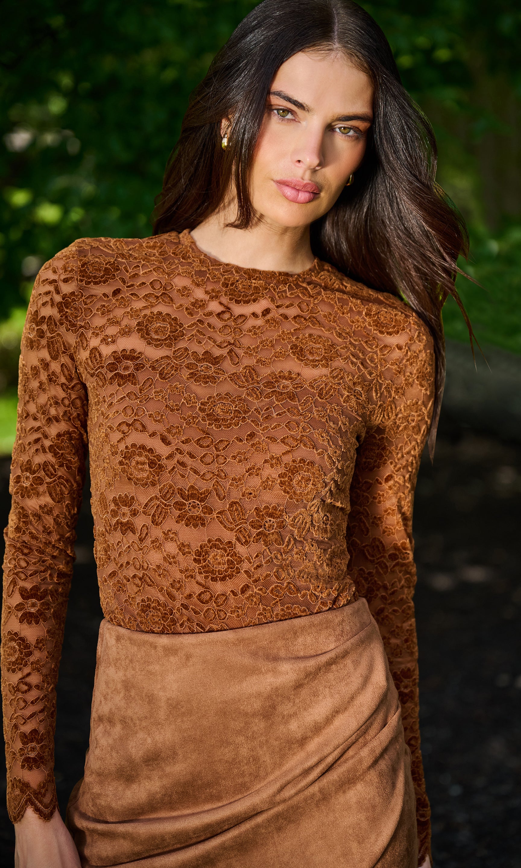 A woman wearing a brown lace long-sleeve top and a matching brown skirt, photographed outdoors with a blurred green background.