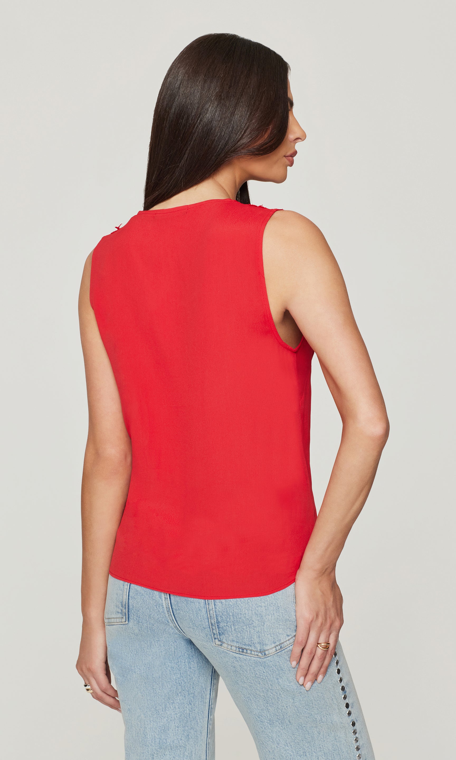 Back view of woman wearing a red sleeveless top and light blue jeans on a plain background