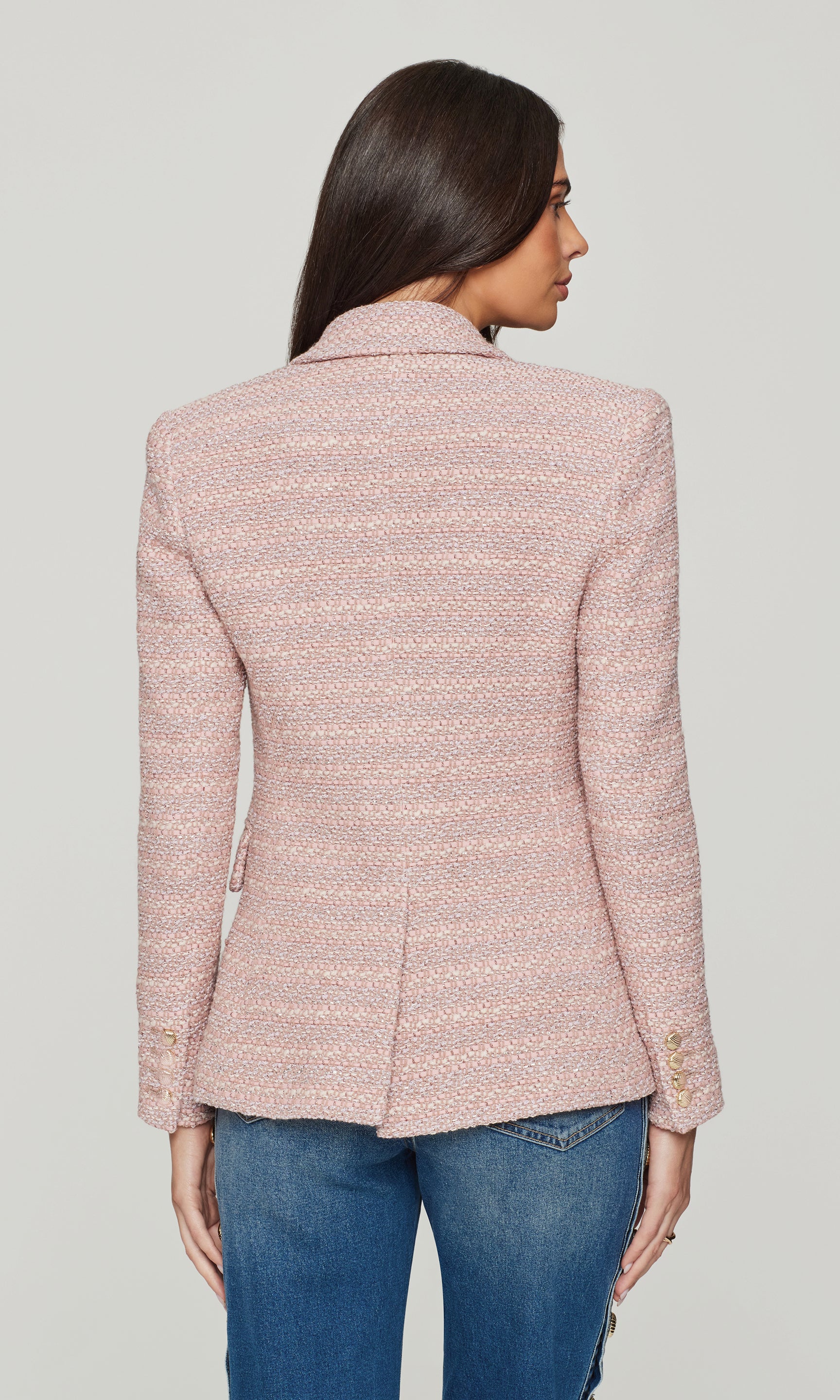 Back view of woman wearing a pink tweed blazer and blue jeans on a light gray background