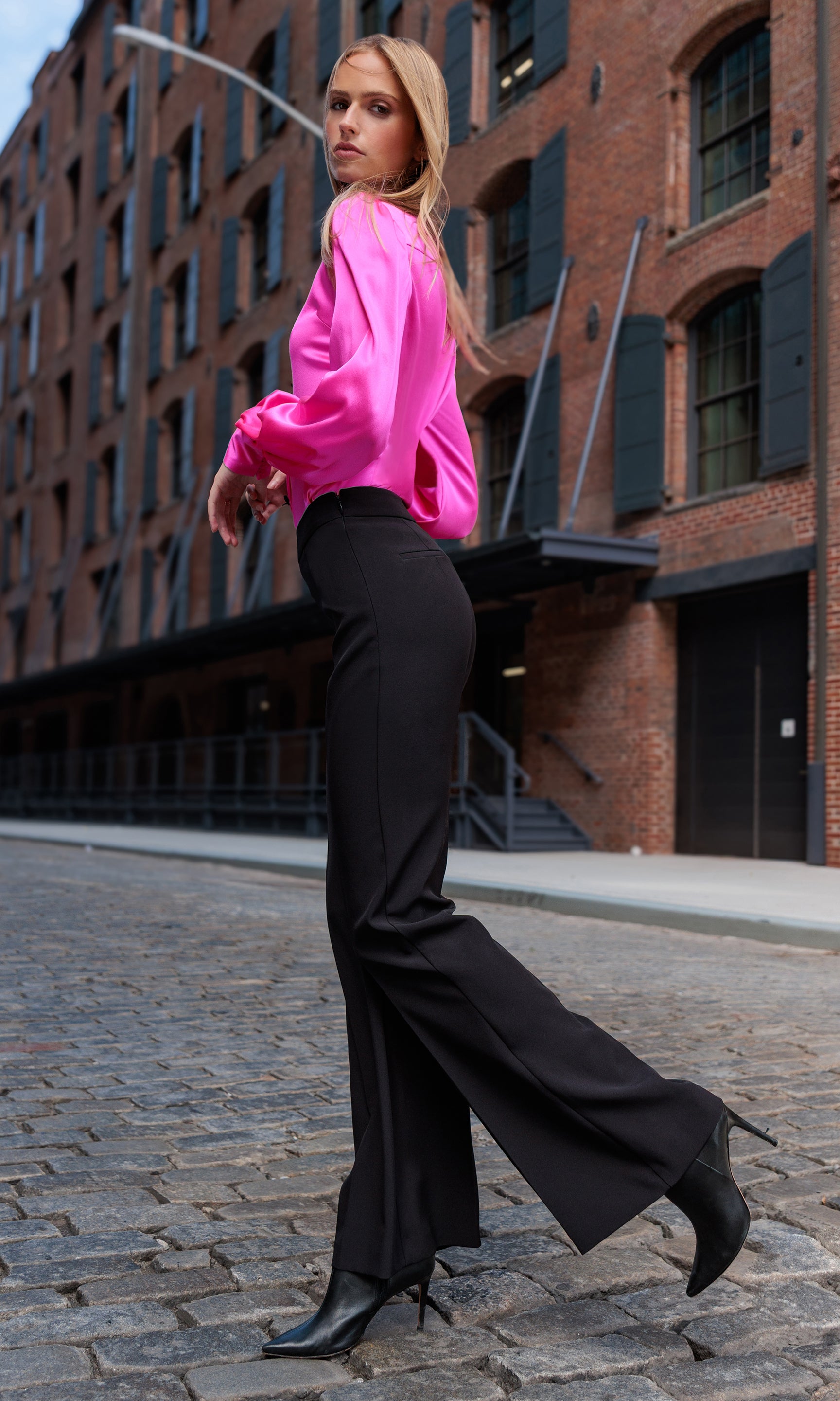 Woman in a bright pink blouse and black pants walking on a cobblestone street.