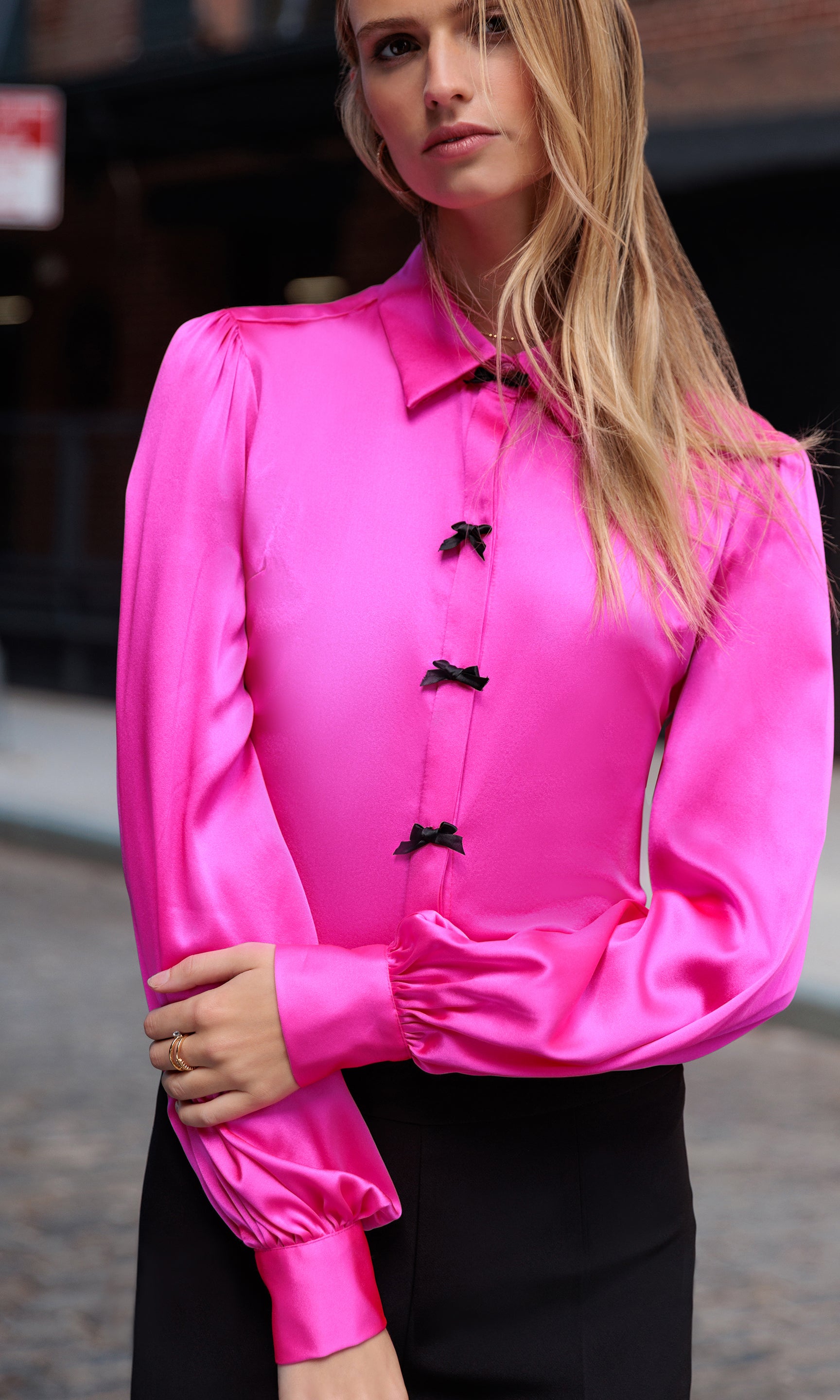 Woman wearing a bright pink blouse with black bows down the front outdoors.