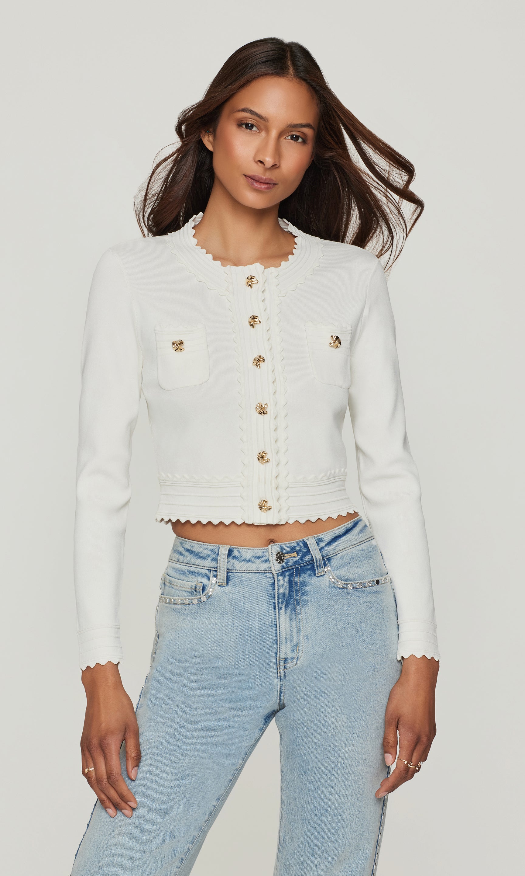 Model wearing a white cardigan with gold buttons and blue jeans on a white background