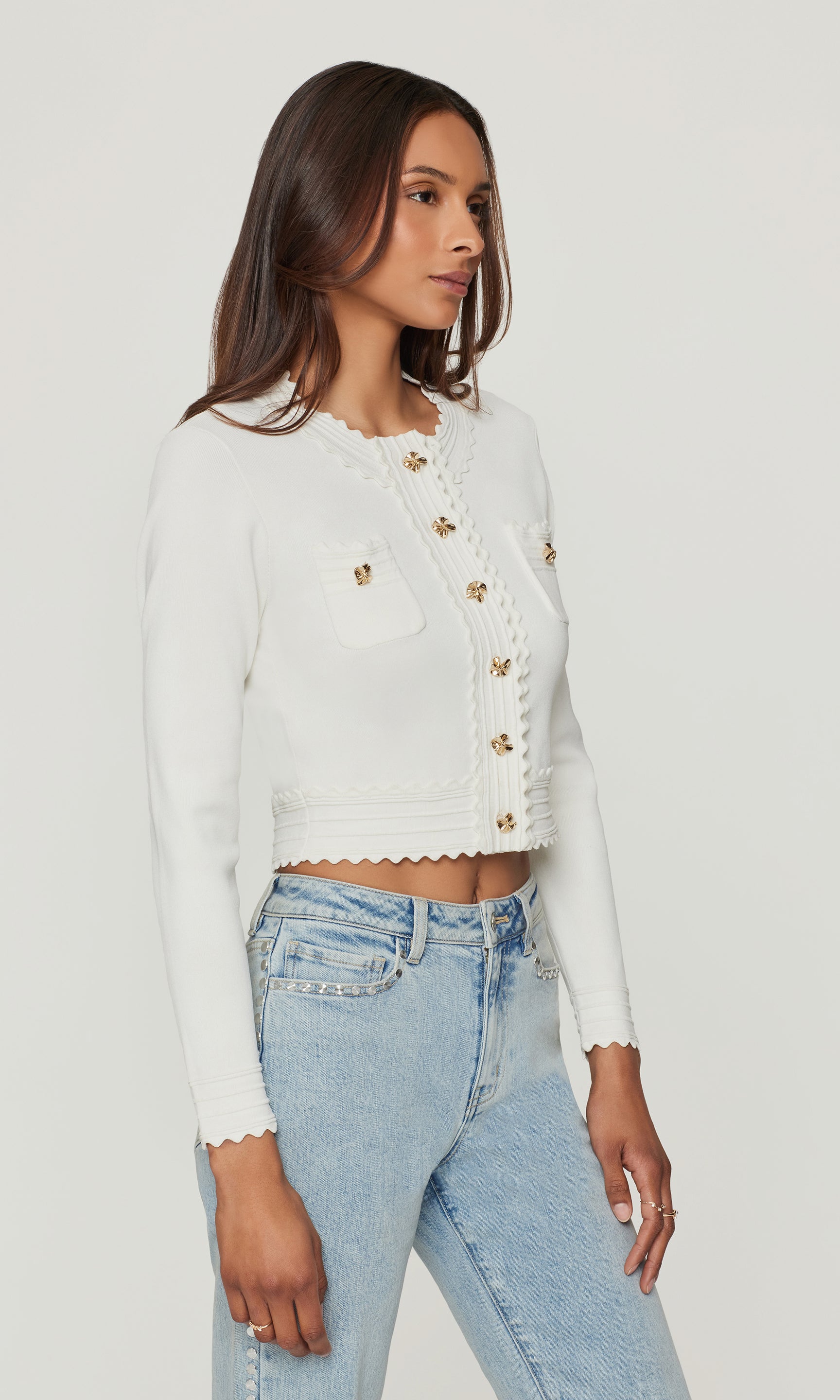 Side view of model wearing a white cardigan with gold buttons and blue jeans on a white background