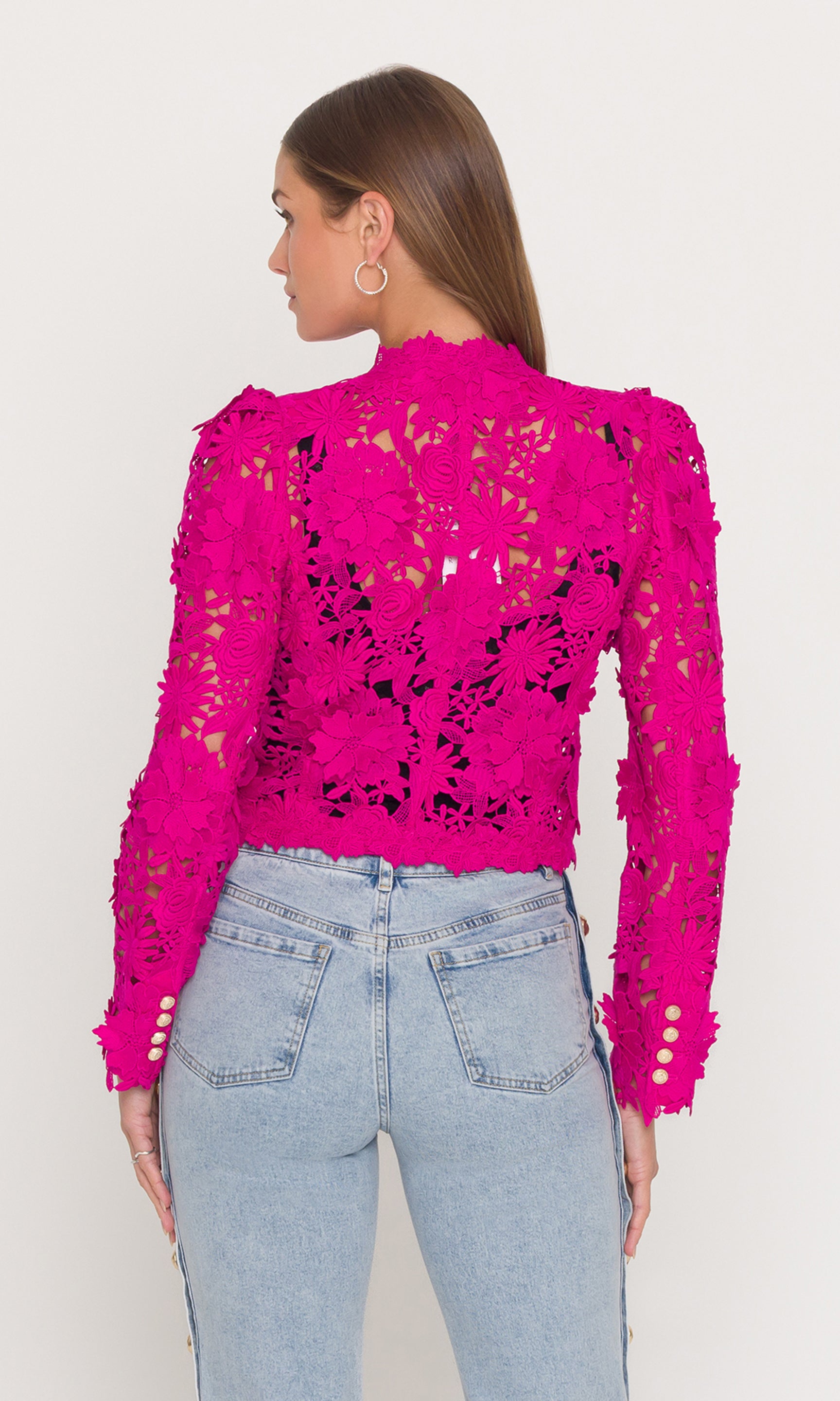 Back view of woman wearing a bright pink lace jacket and light blue jeans on a plain background
