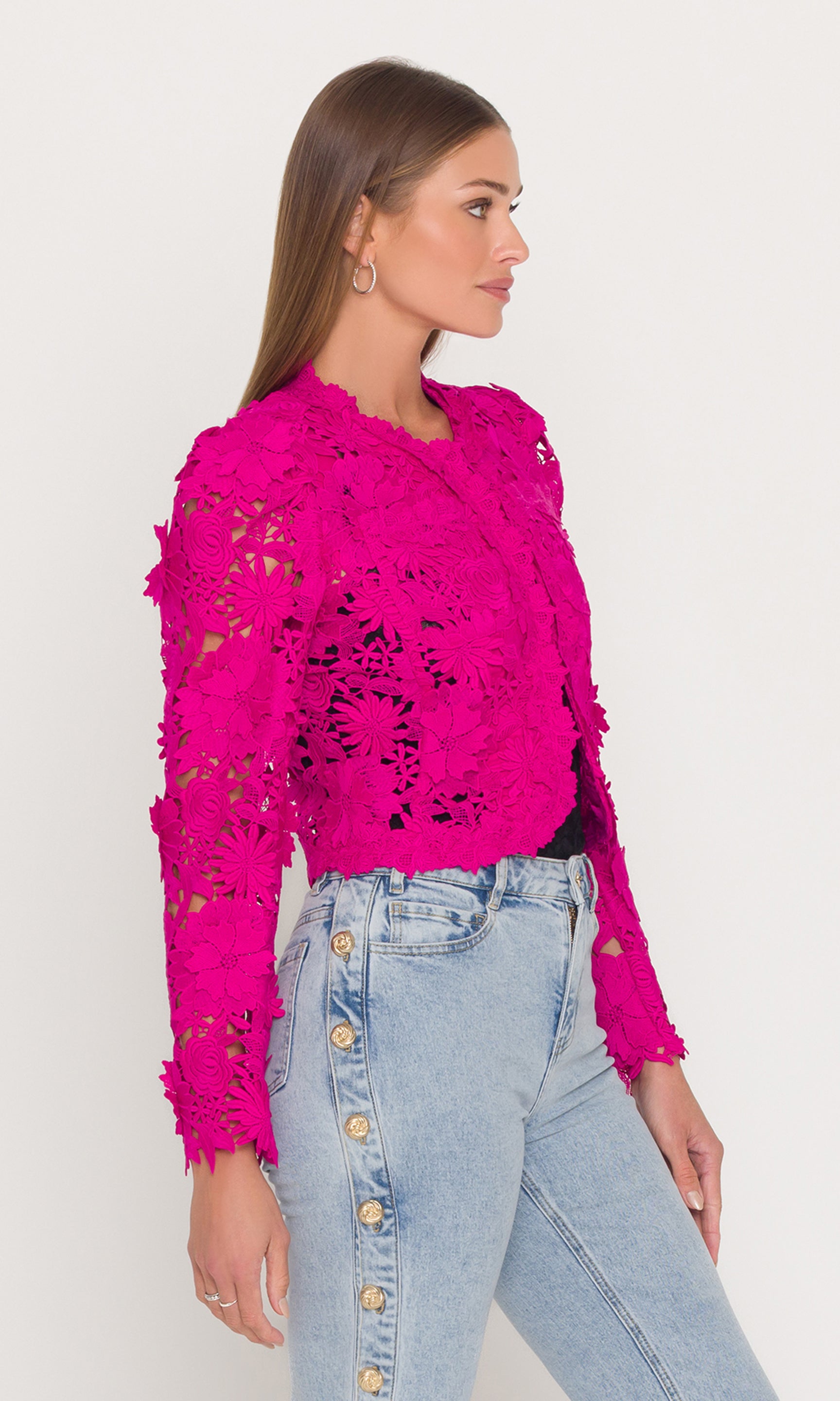 Side view of woman wearing a bright pink lace jacket and light blue jeans against a white background