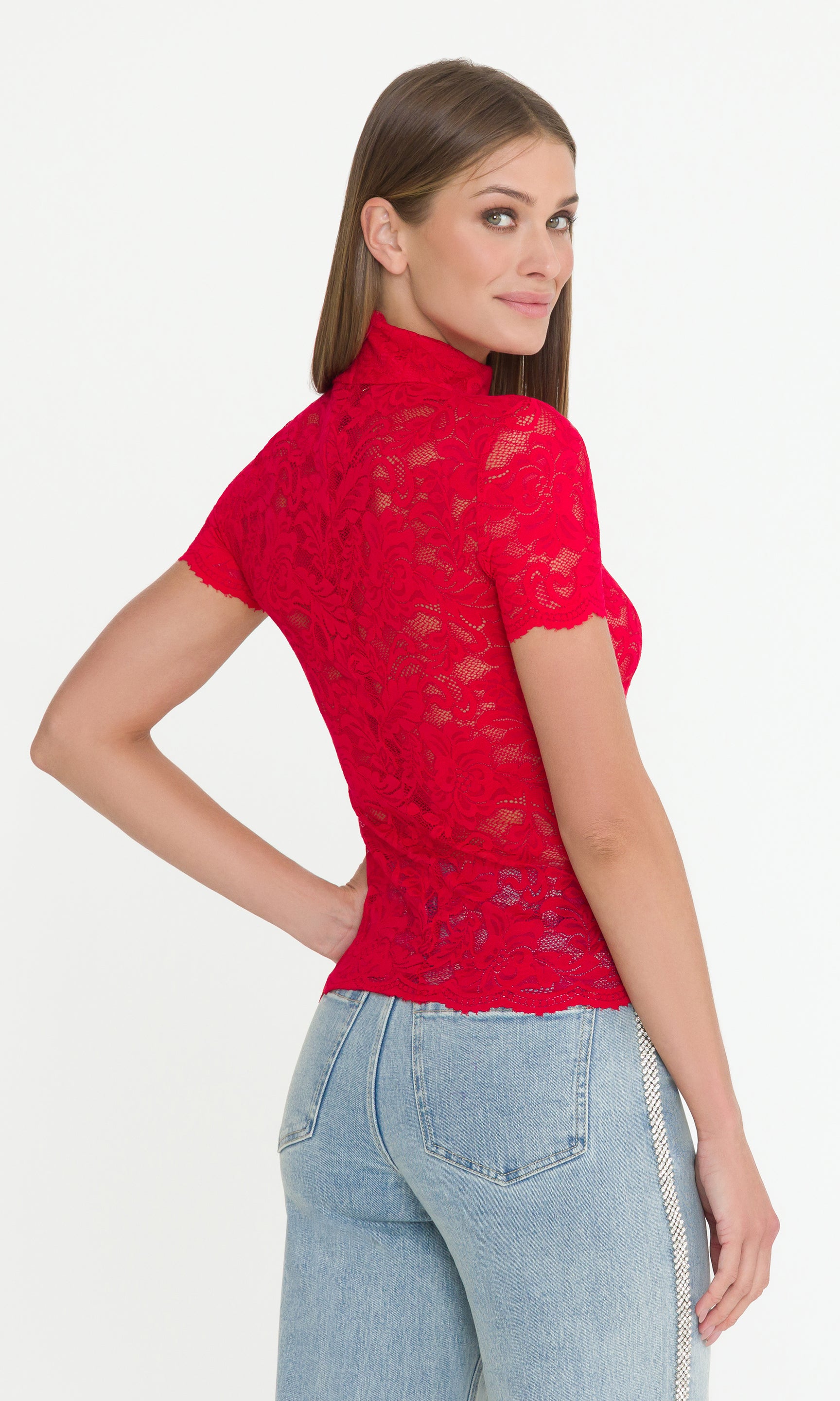 Back view of model looking over her shoulder wearing a red lace top and light blue jeans on a white background