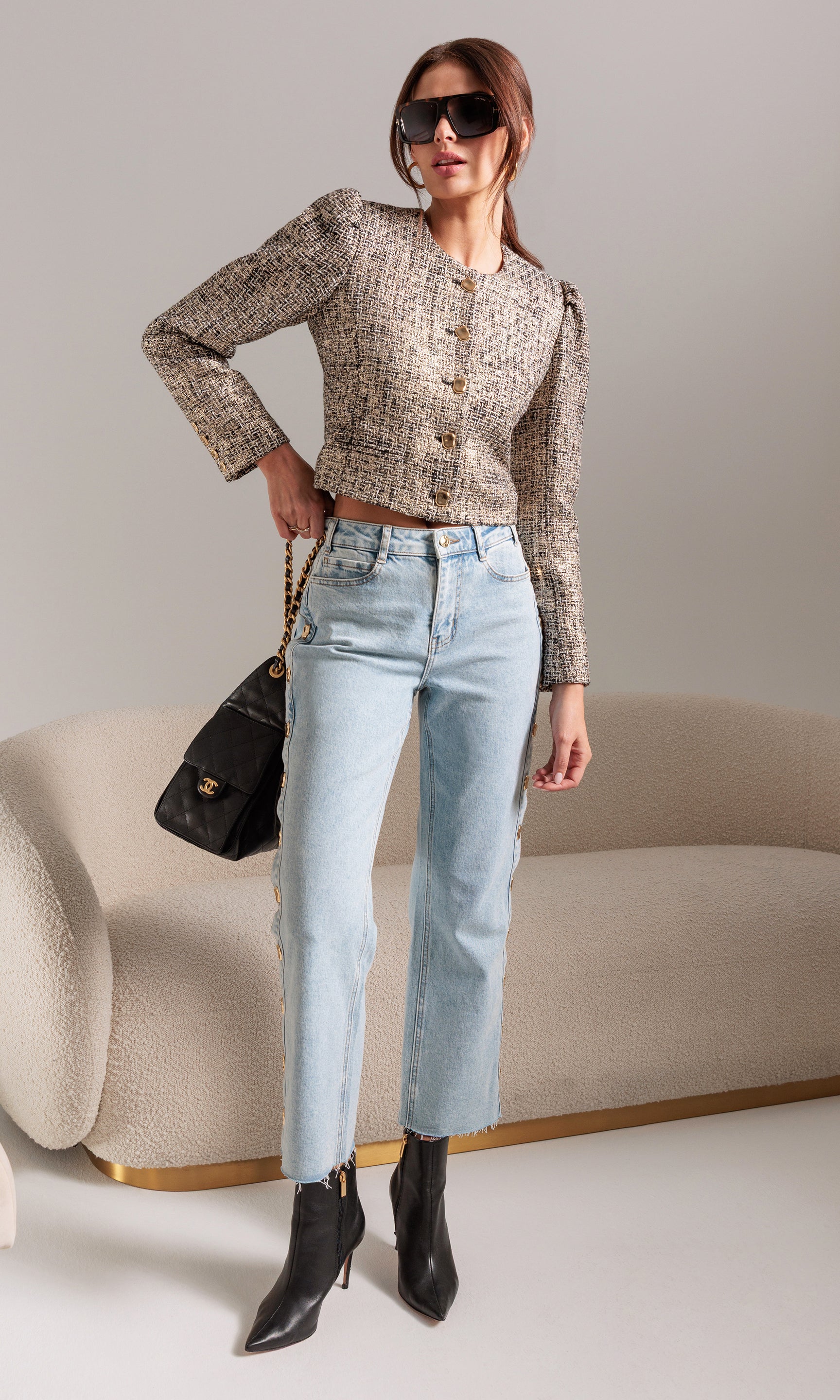 Woman in a stylish outfit with a textured top, light blue jeans, black boots, and sunglasses, holding a black handbag.