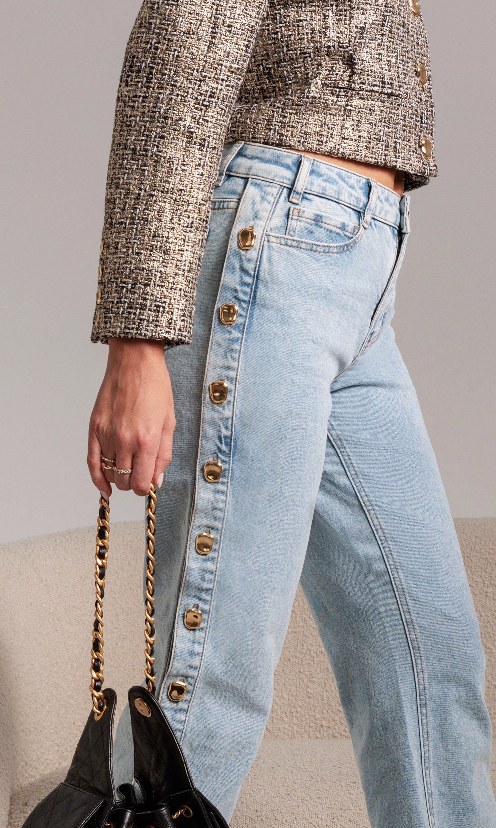 Person wearing light blue jeans with gold buttons, holding a black handbag with gold chain strap.