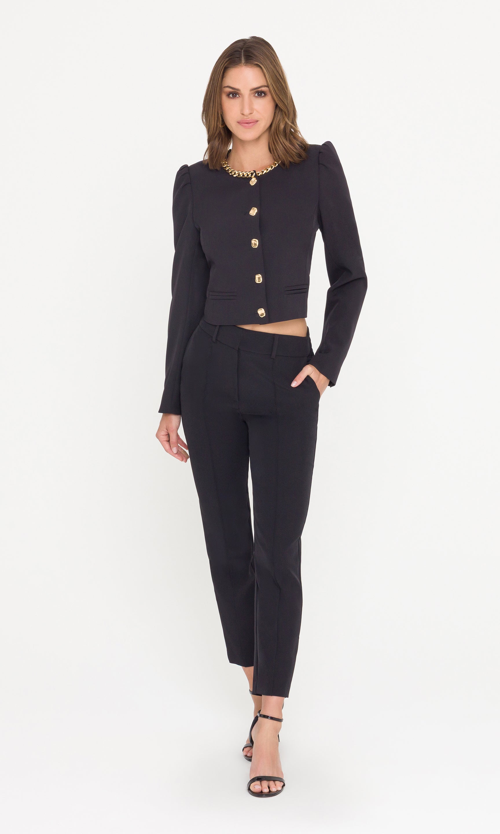 Full body image of model wearing a black cropped jacket with a gold chain neckline and gold buttons and black pants