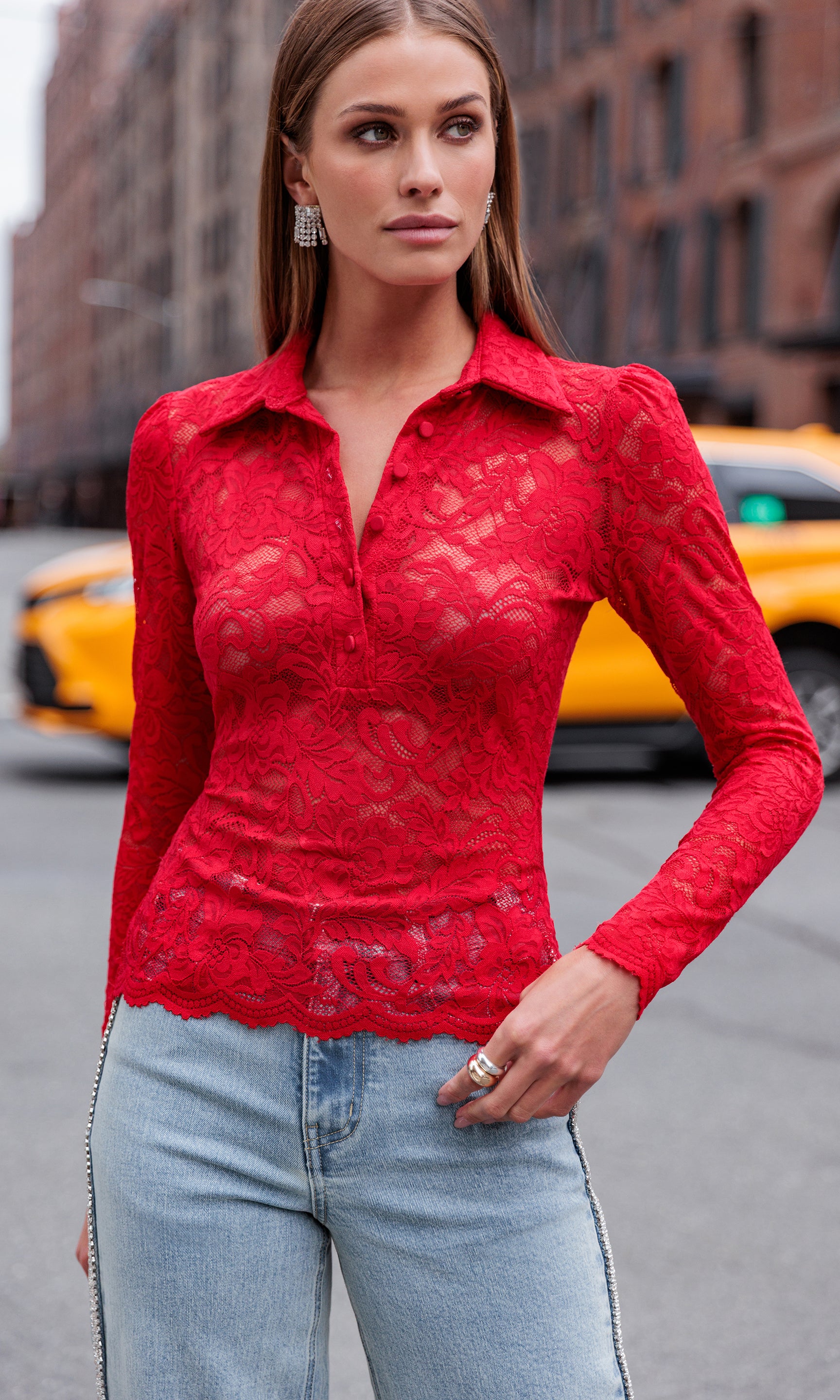 Woman wearing a red lace top and light blue jeans standing on a city street.
