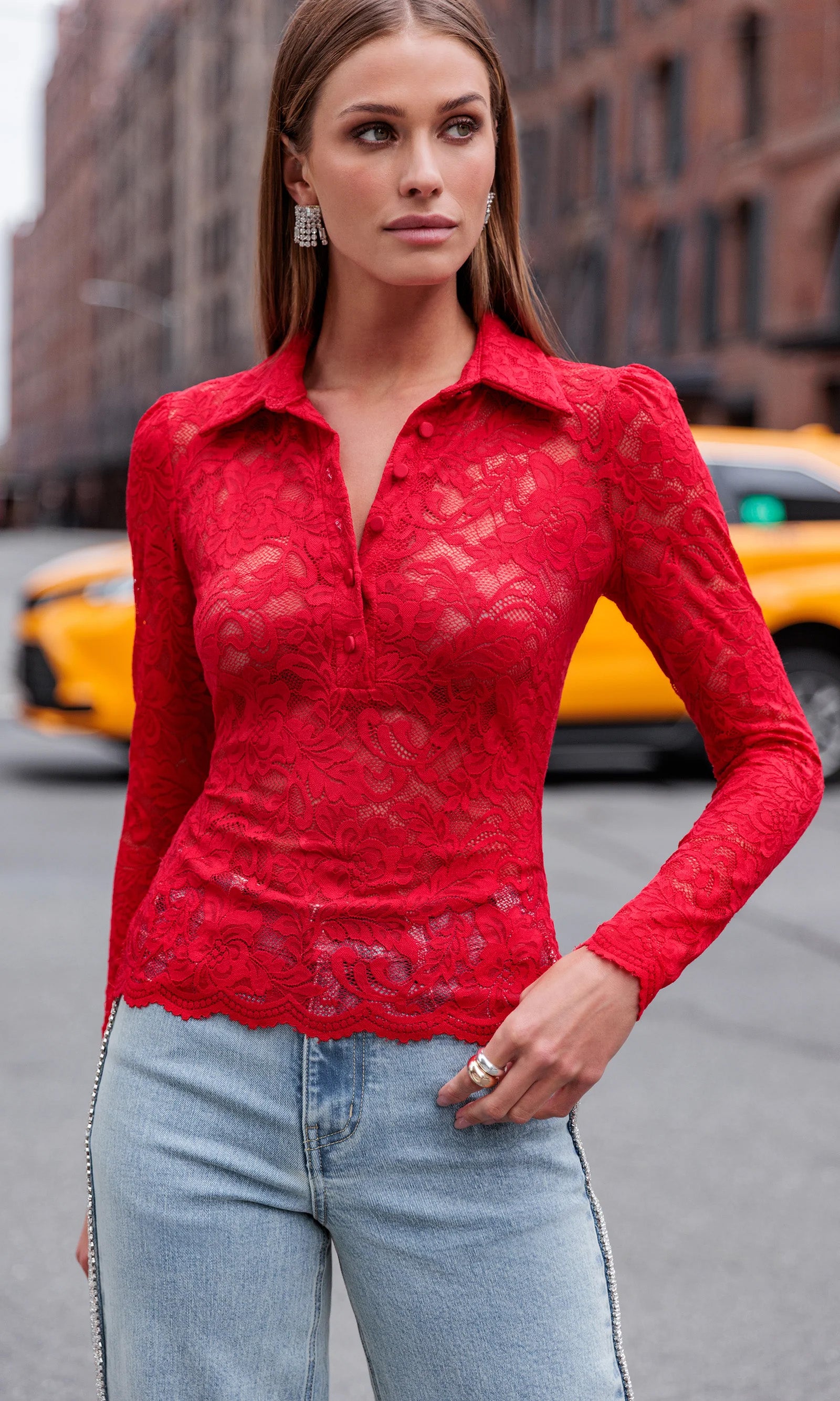 Woman wearing a red lace top and light blue jeans standing on a city street.