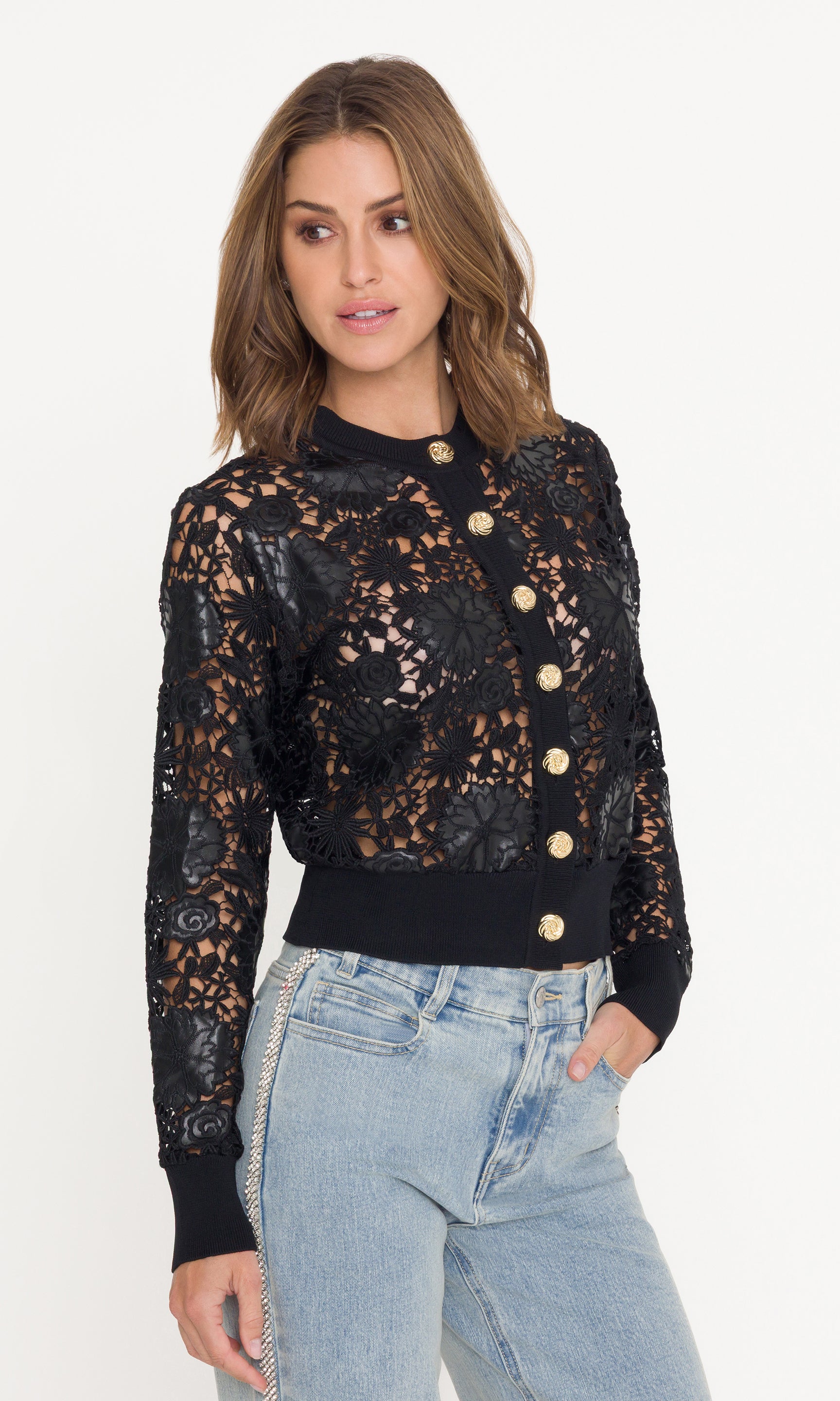Woman wearing a black lace cardigan with gold buttons and light blue jeans on a white background
