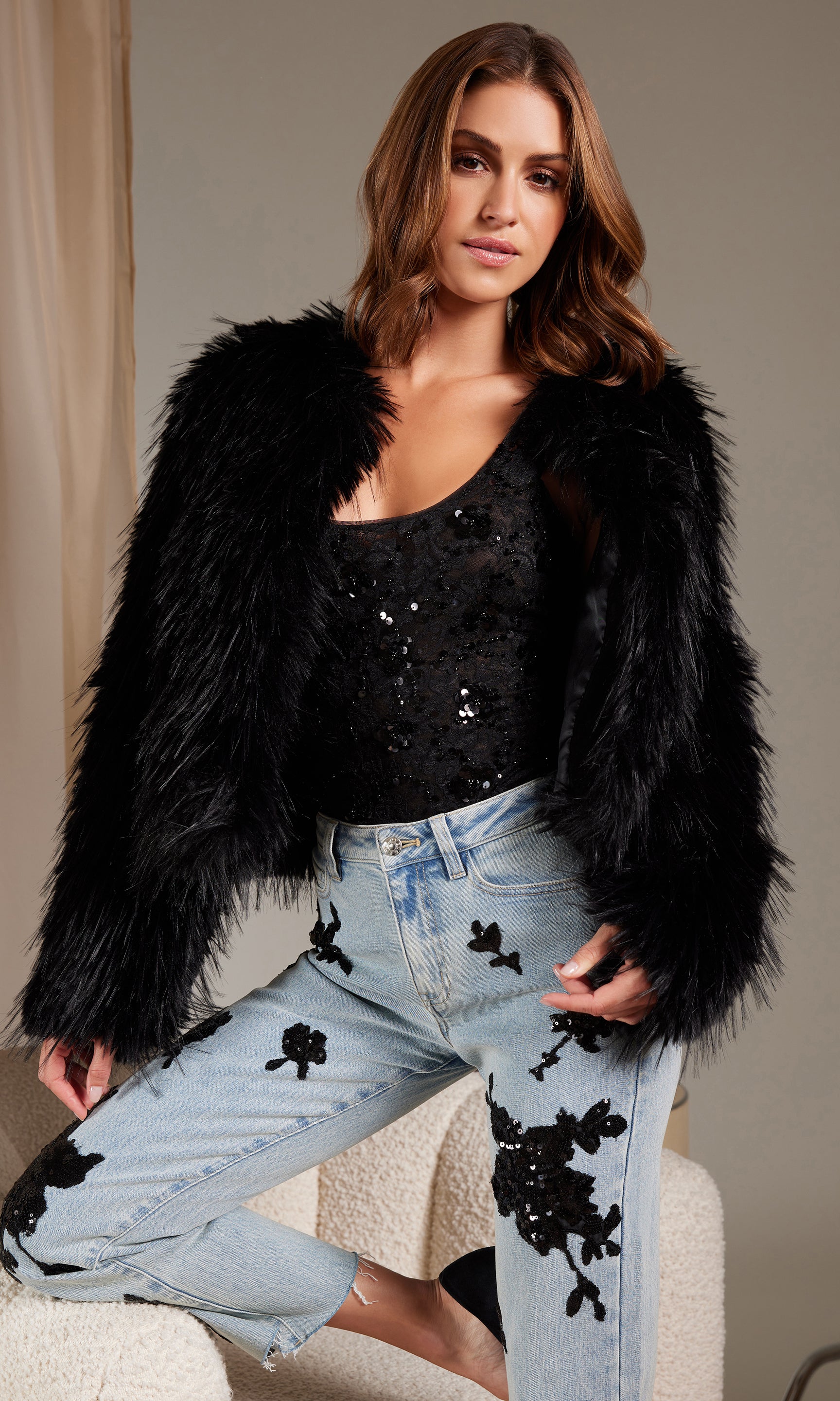 Woman wearing a black fur coat, black sequin top, and light blue jeans with black floral patterns.