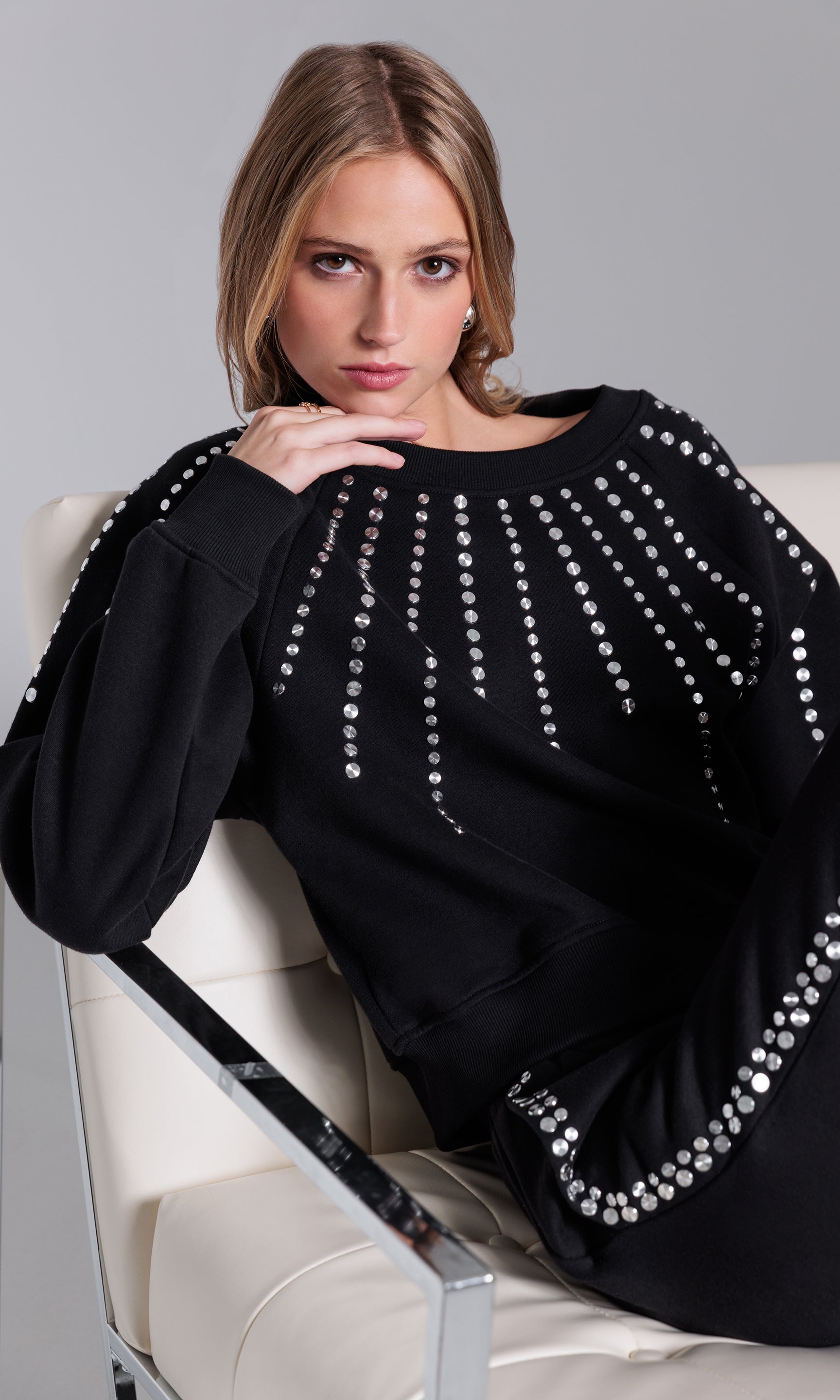Woman wearing a black sweater with silver embellishments sitting on a white chair.
