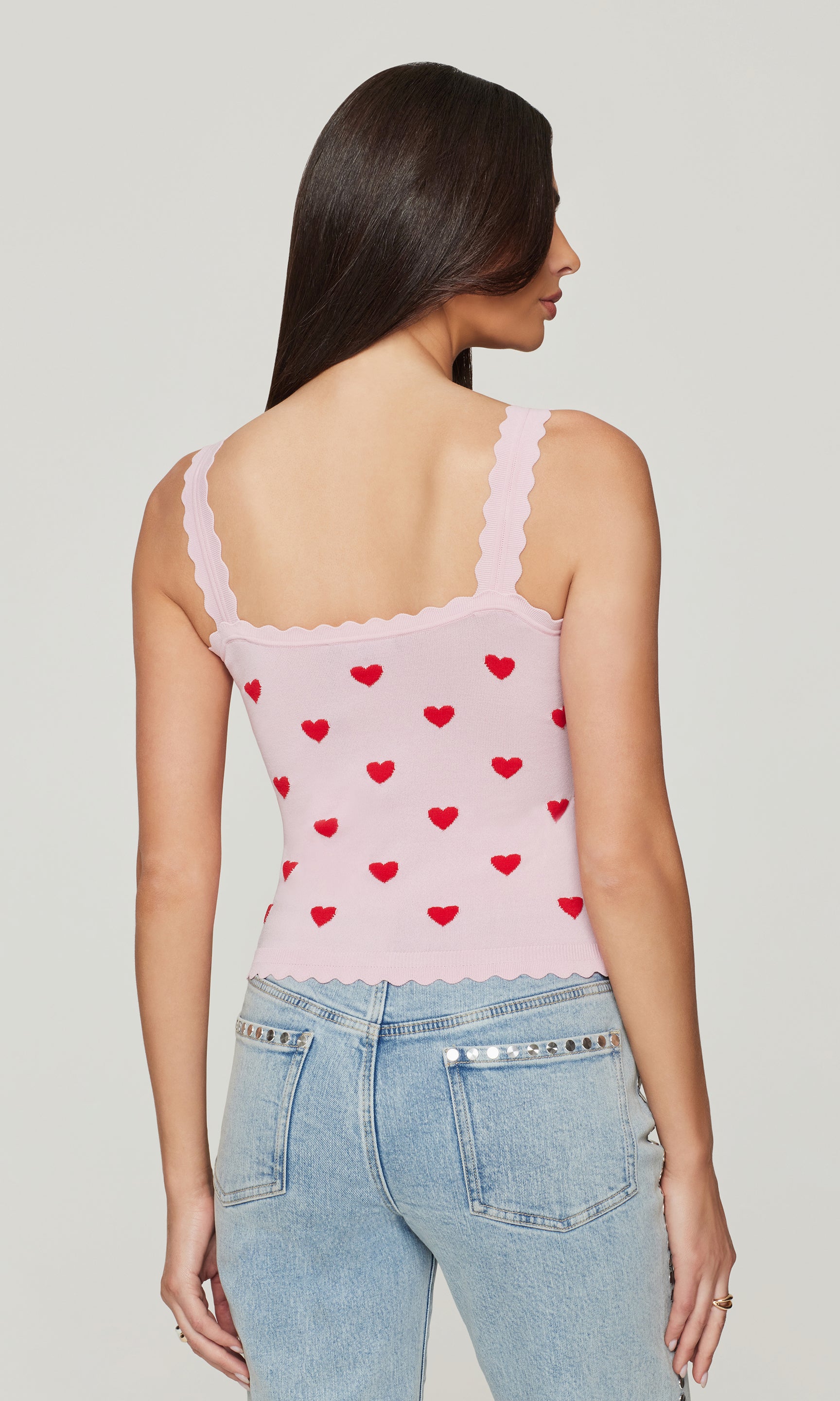 Back view of woman wearing a pink tank top with red heart patterns and light blue jeans on a plain background