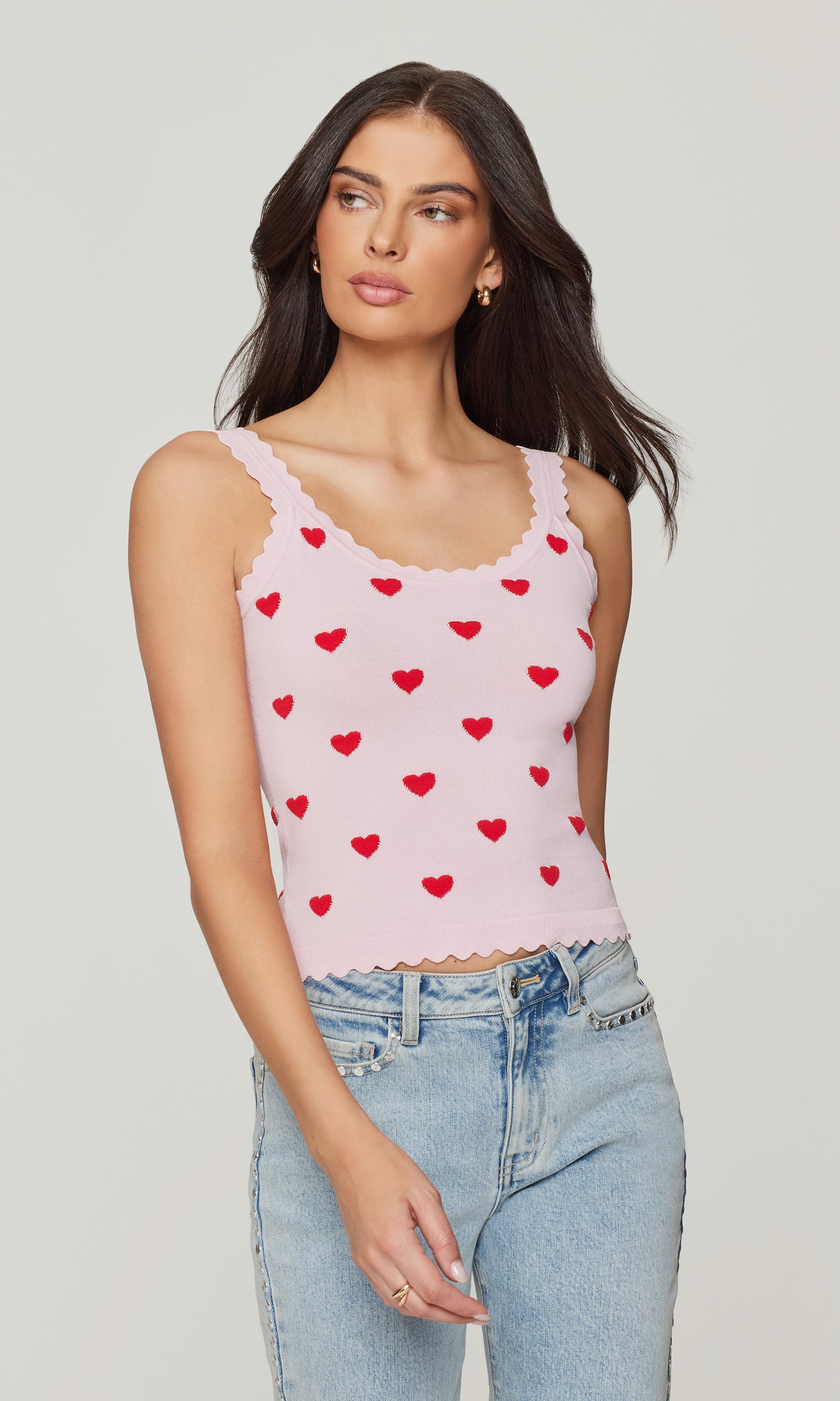 Woman wearing a pink tank top with red heart patterns and light blue jeans on a plain background