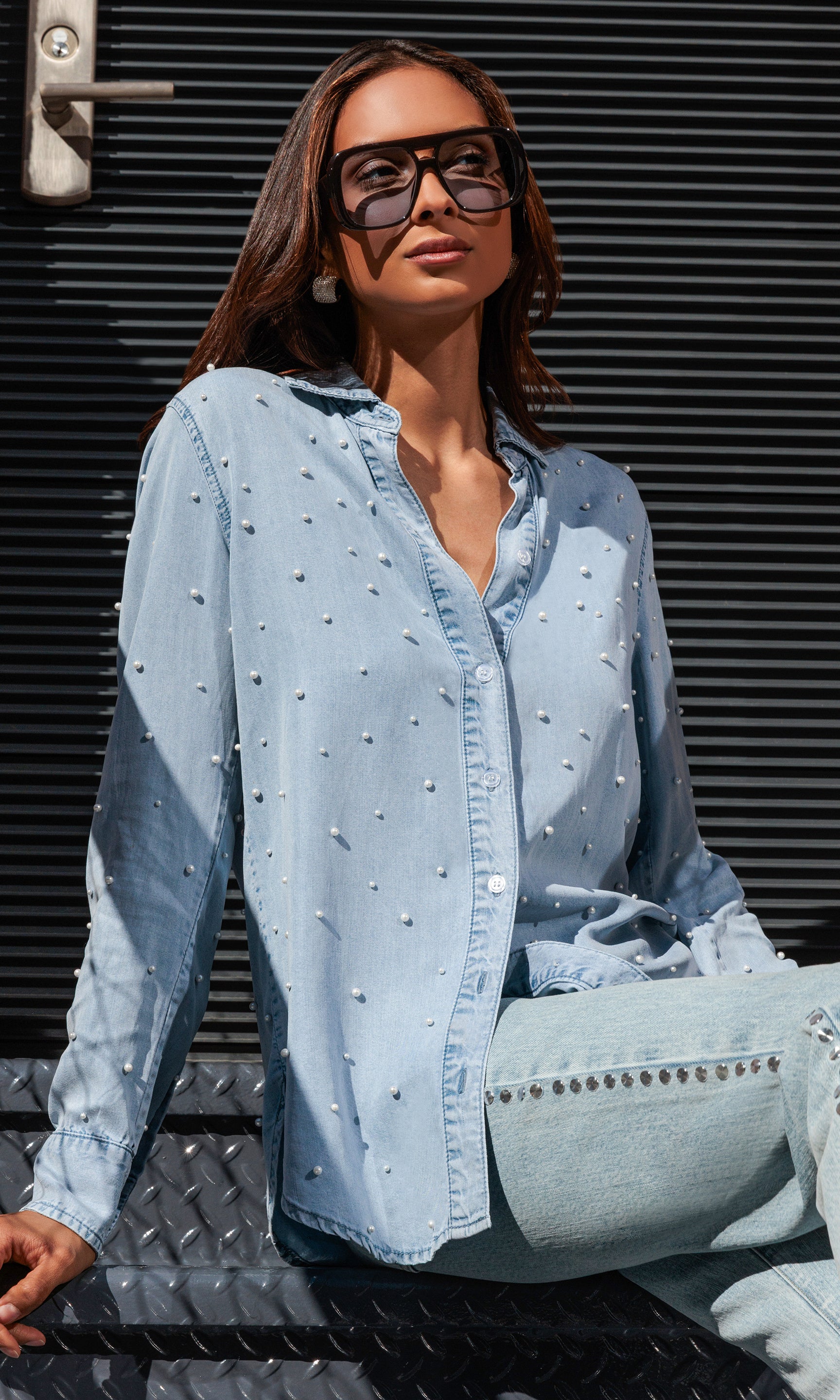 Woman wearing a light blue denim shirt with pearls, sitting against a metallic background.