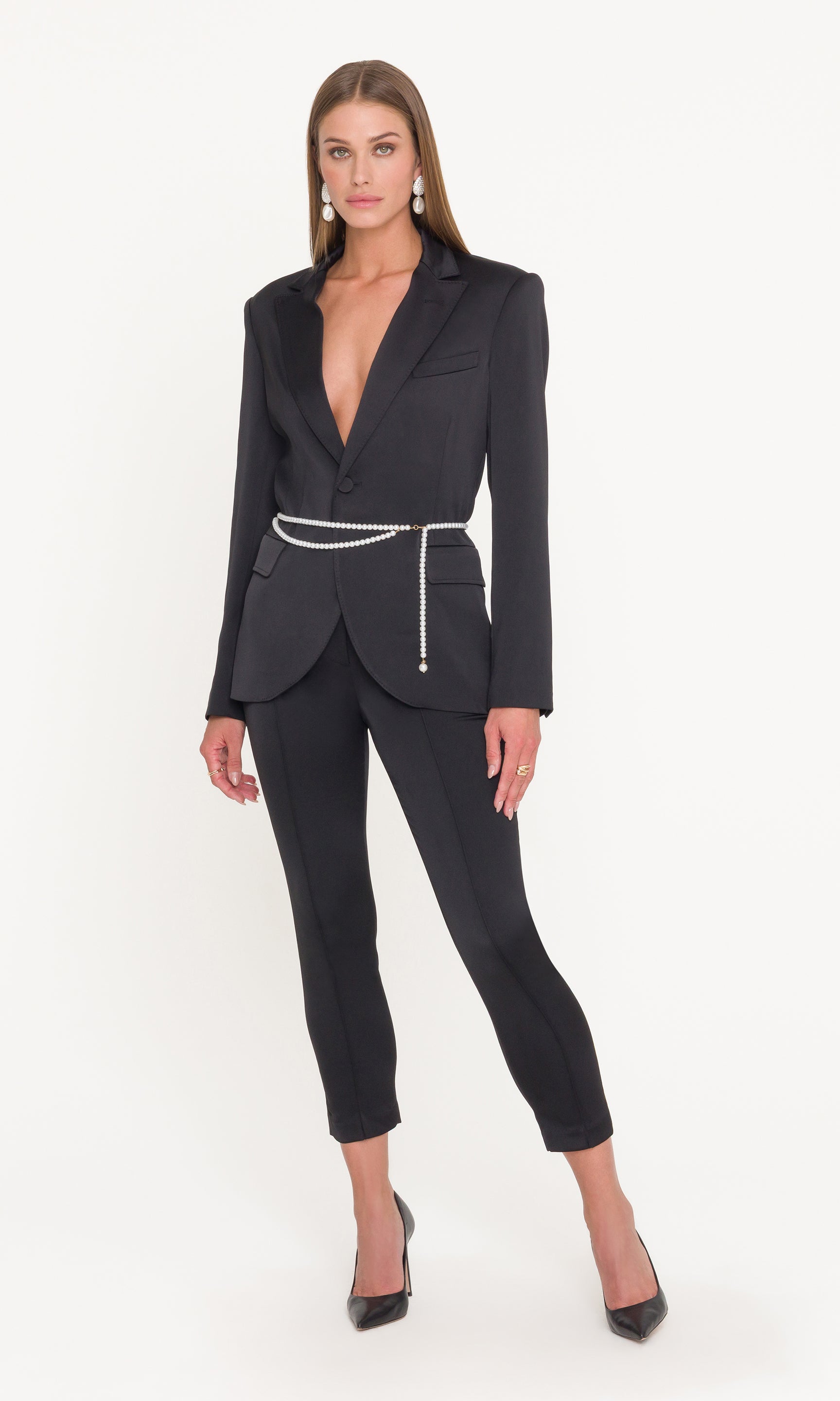 Full body image of model wearing a black satin suit with a pearl belt on a white background