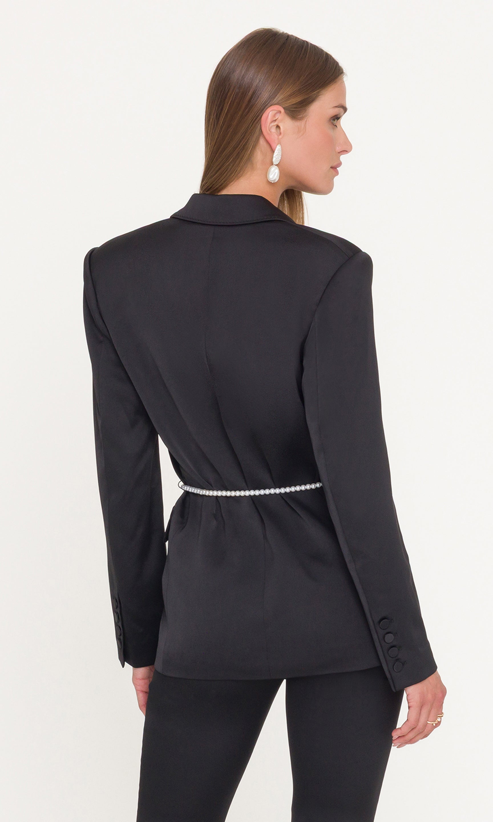 Back view of model wearing a black suit with a pearl belt on a white background