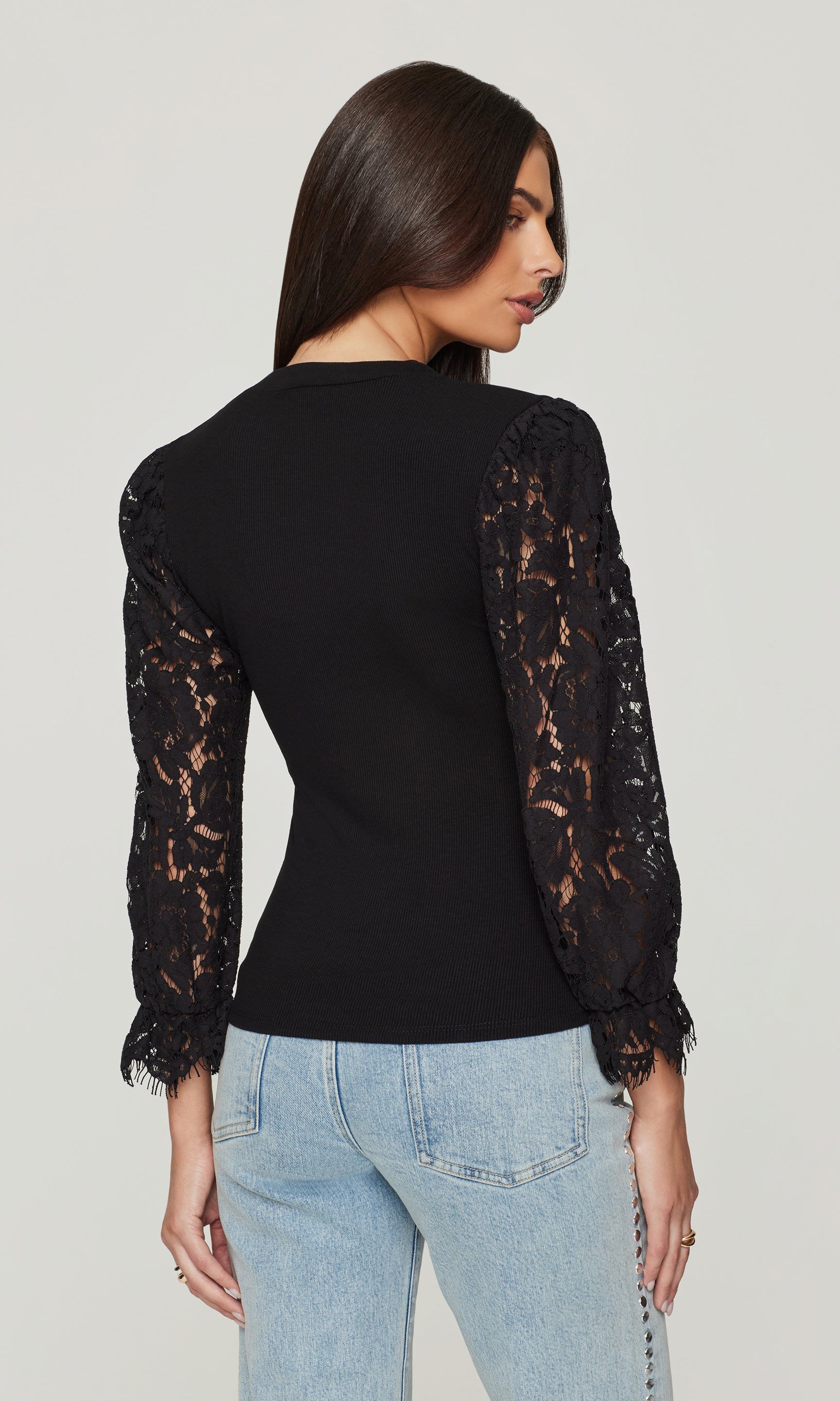 Back view of woman wearing a black top with lace sleeves and light blue jeans on a plain background