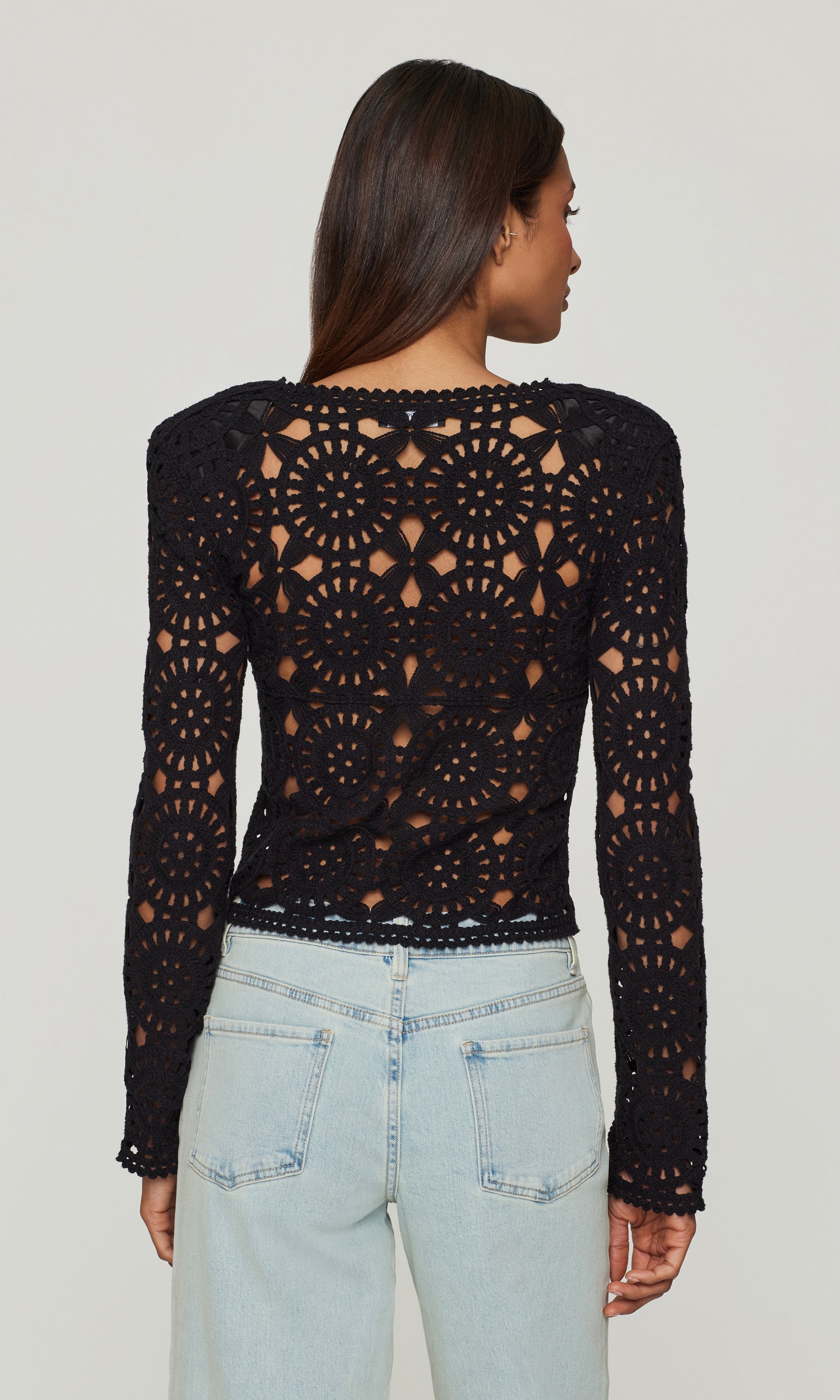 Back view of model wearing a black crochet long-sleeve top and light blue jeans on a plain background