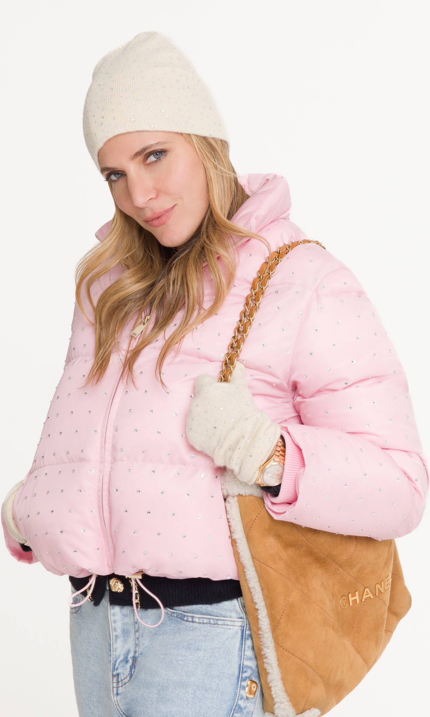 Woman wearing a pink puffer jacket with a beige beanie and gloves, holding a brown bag.