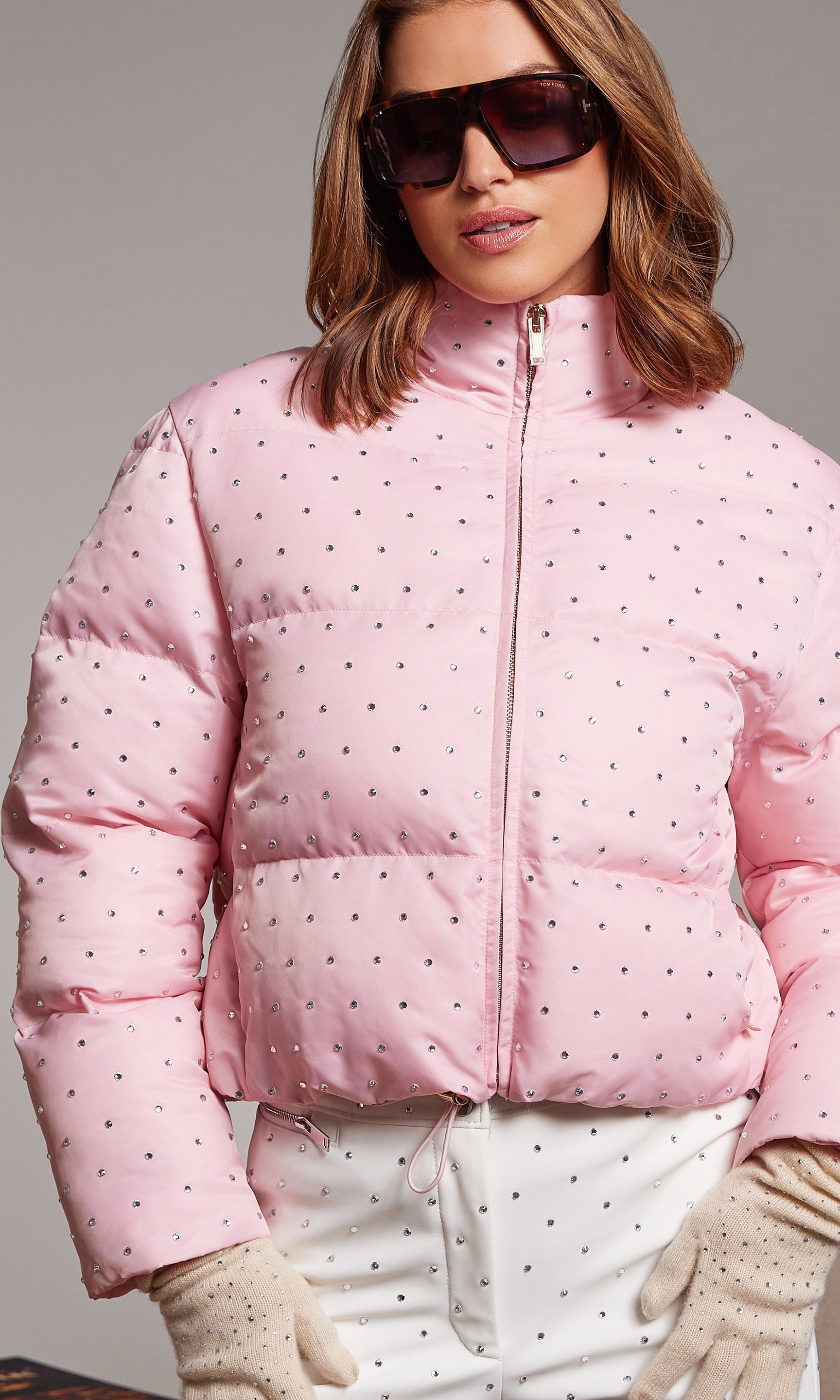 Woman wearing a pink crystal puffer jacket with sunglasses and gloves on a gray background