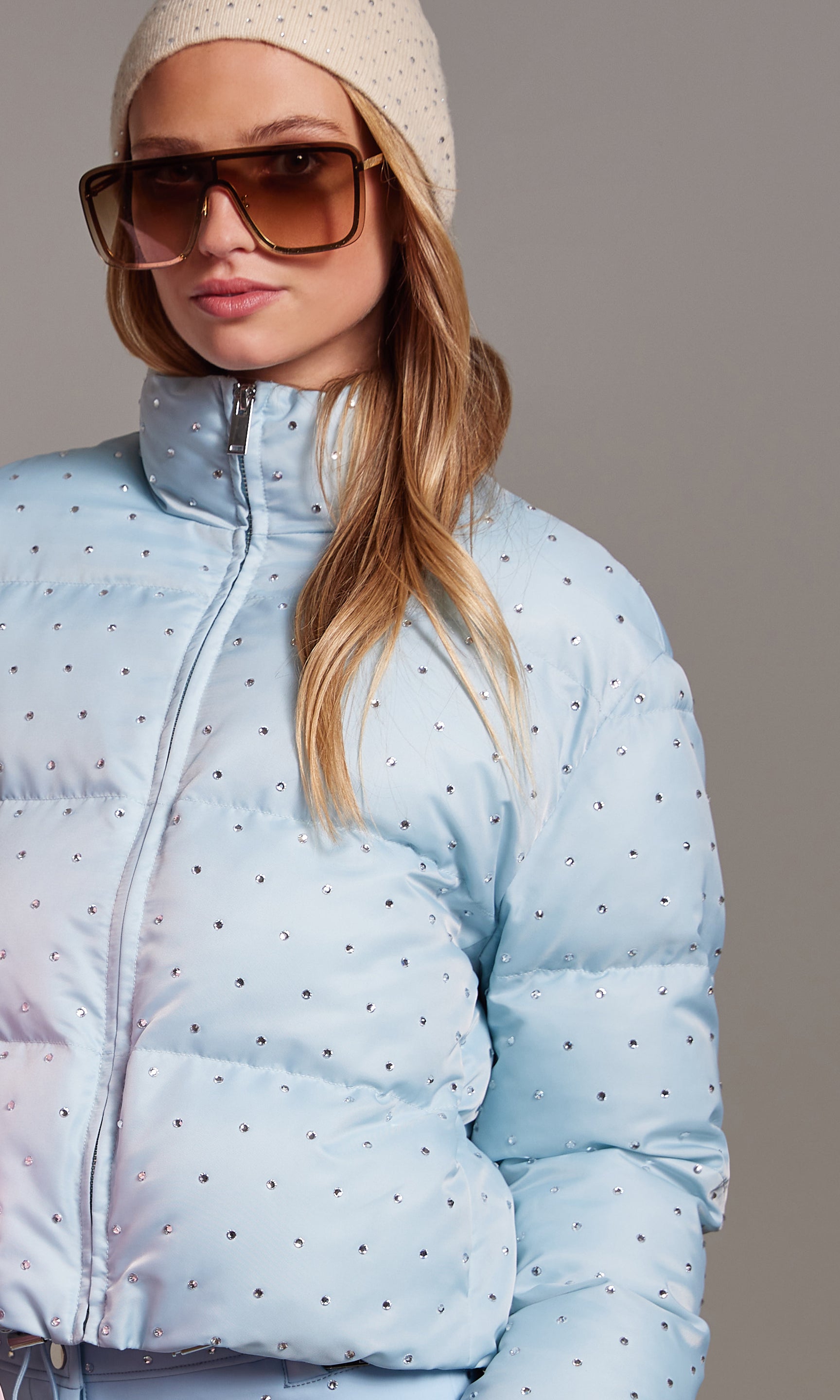 Person wearing a light blue puffer jacket with a subtle pattern, sunglasses, and a beige beanie against a gray background.