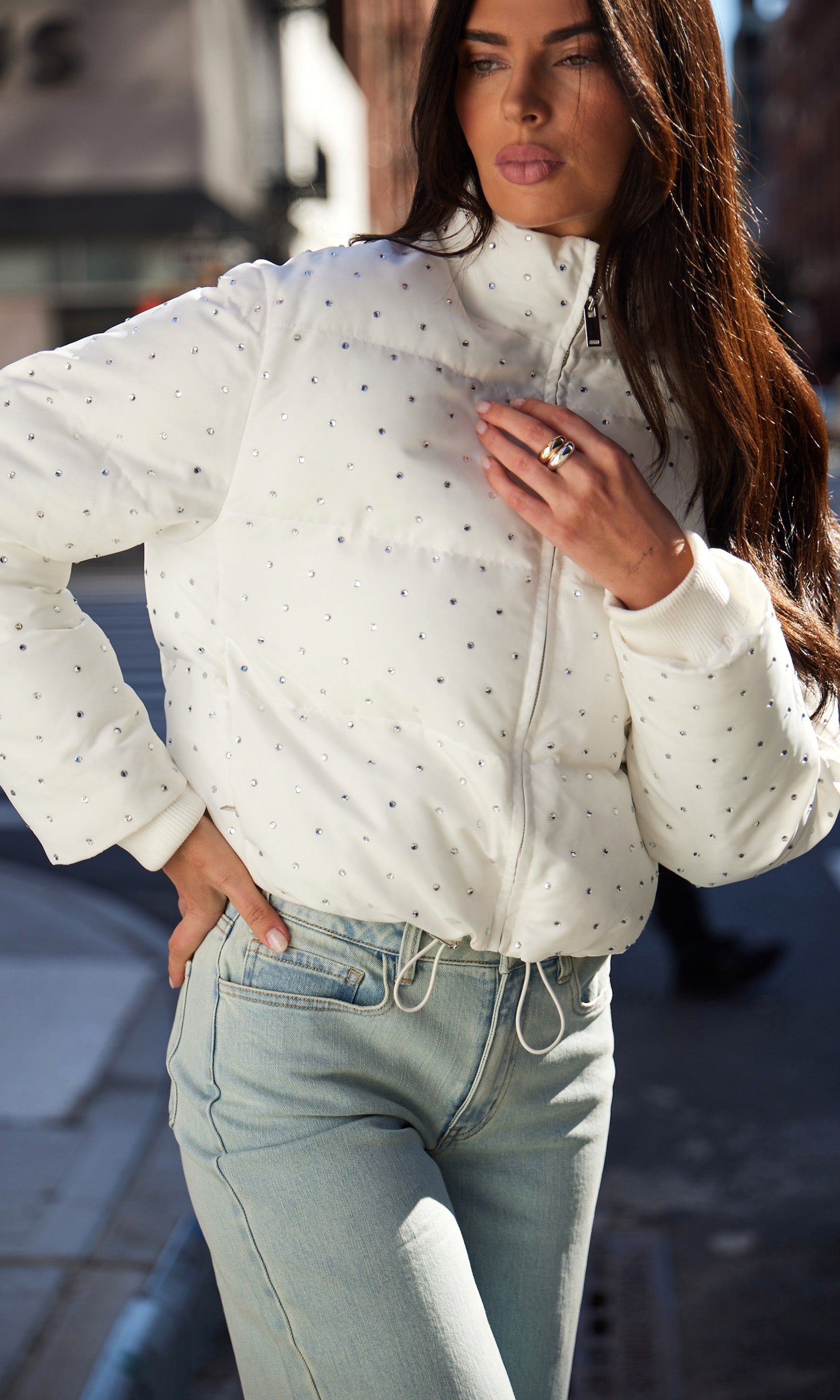 Woman wearing a white crystal covered jacket and light blue jeans on a blurred street background