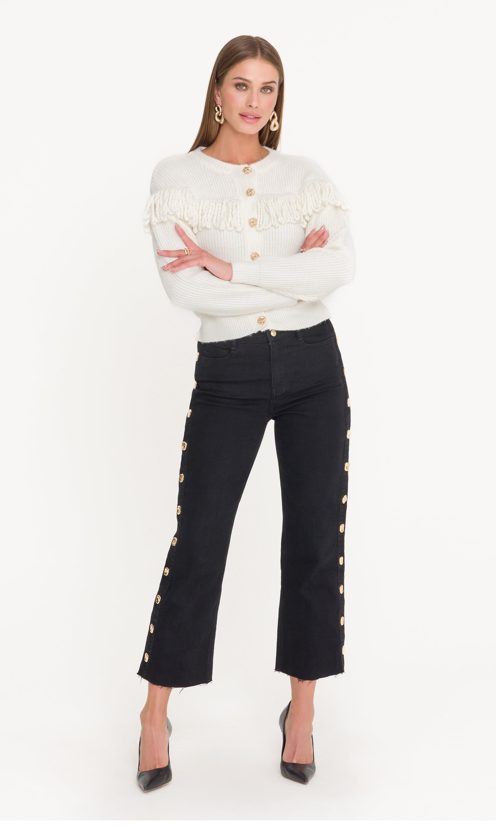 Woman wearing a white sweater with fringe detail and black pants with gold buttons on a white background