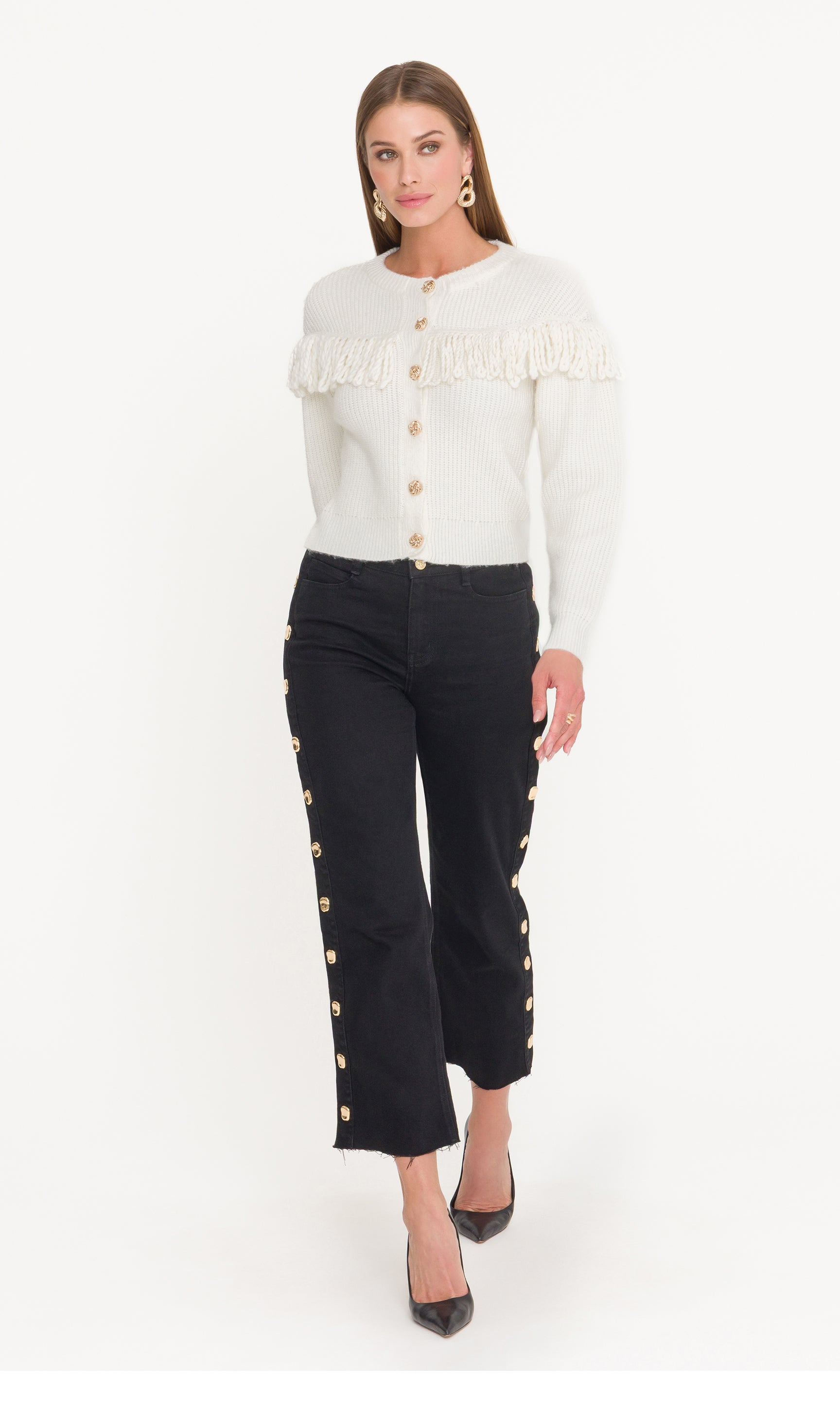 Woman wearing a white sweater with fringe details and black pants with gold buttons on a white background