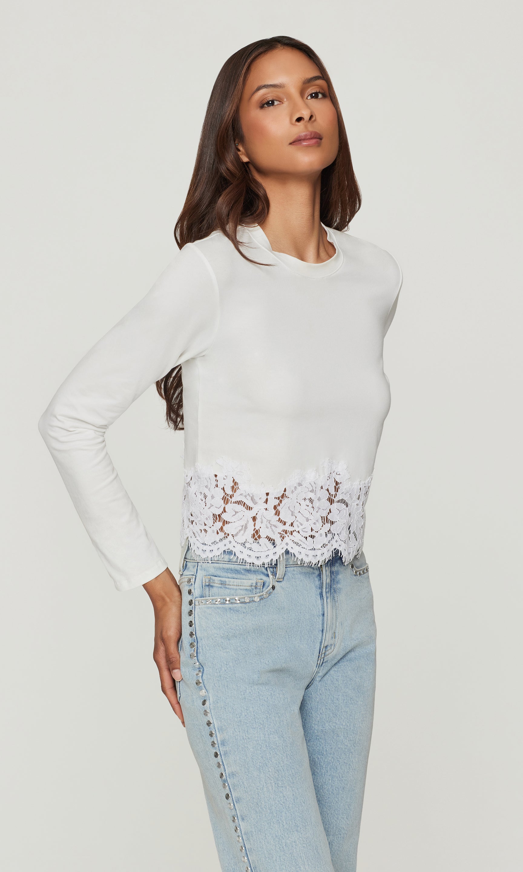 Woman wearing a white long-sleeve top with lace detail and light blue jeans on a white background