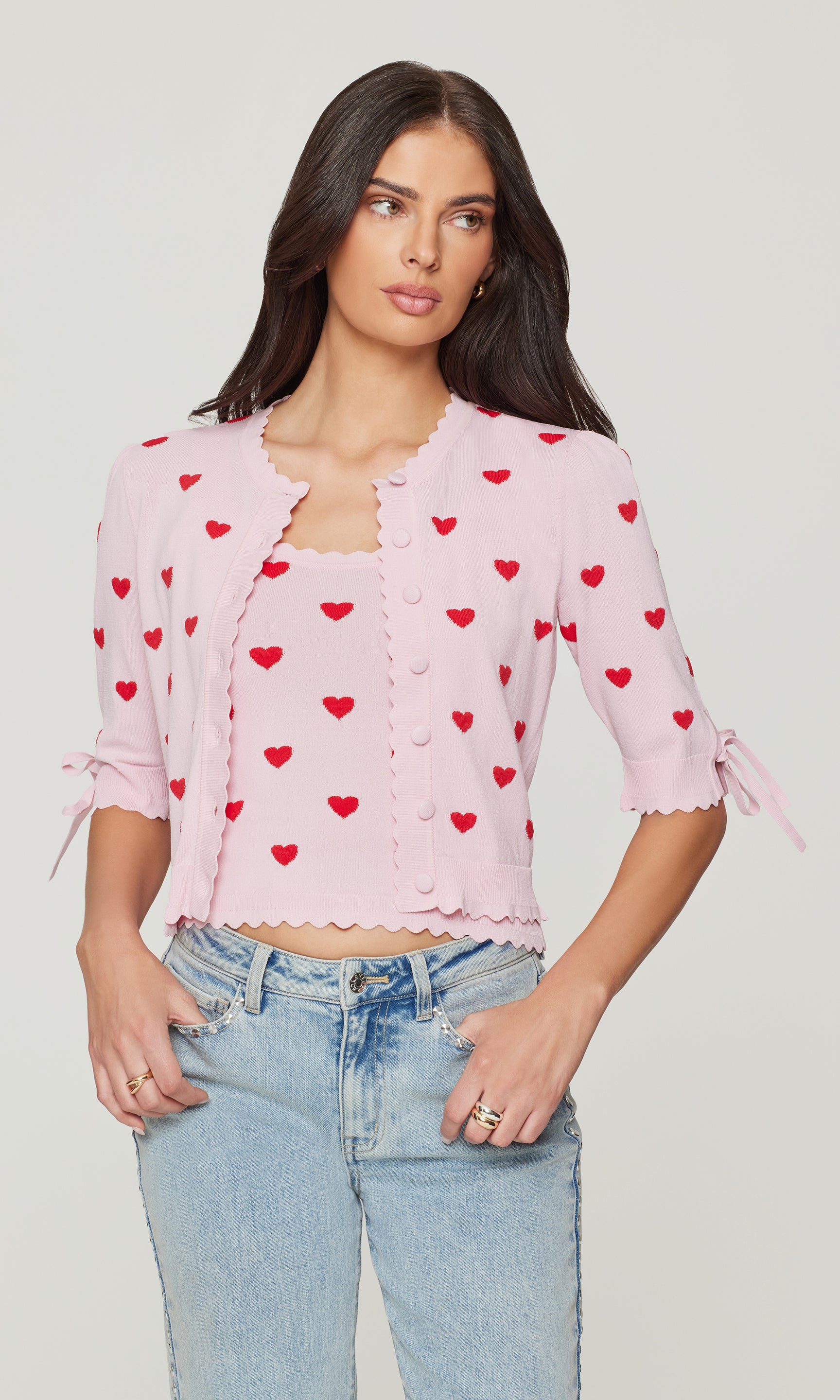 Woman wearing a pink blouse with red heart patterns and light blue jeans on a plain background