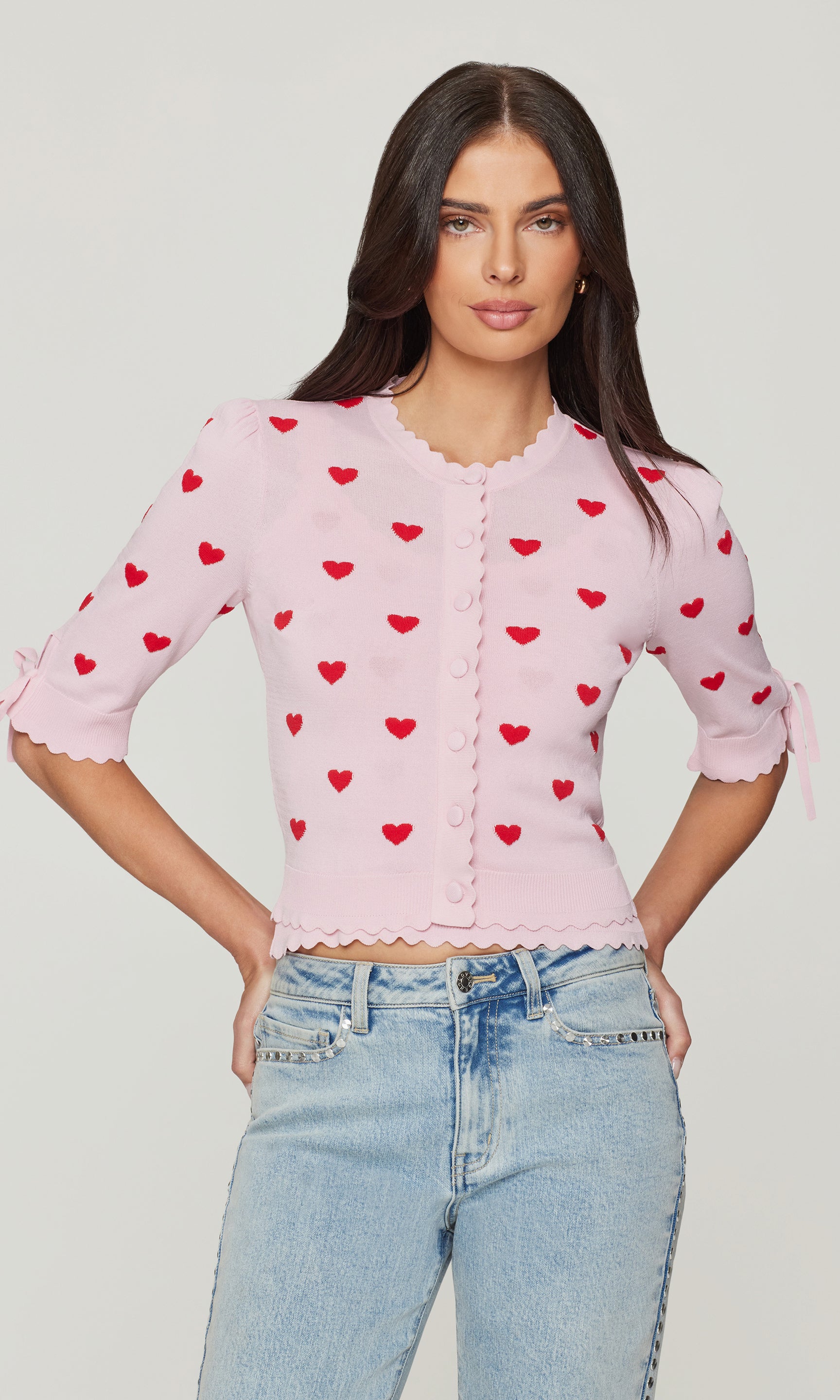 Woman wearing a pink cardigan with red heart patterns and light blue jeans on a plain background