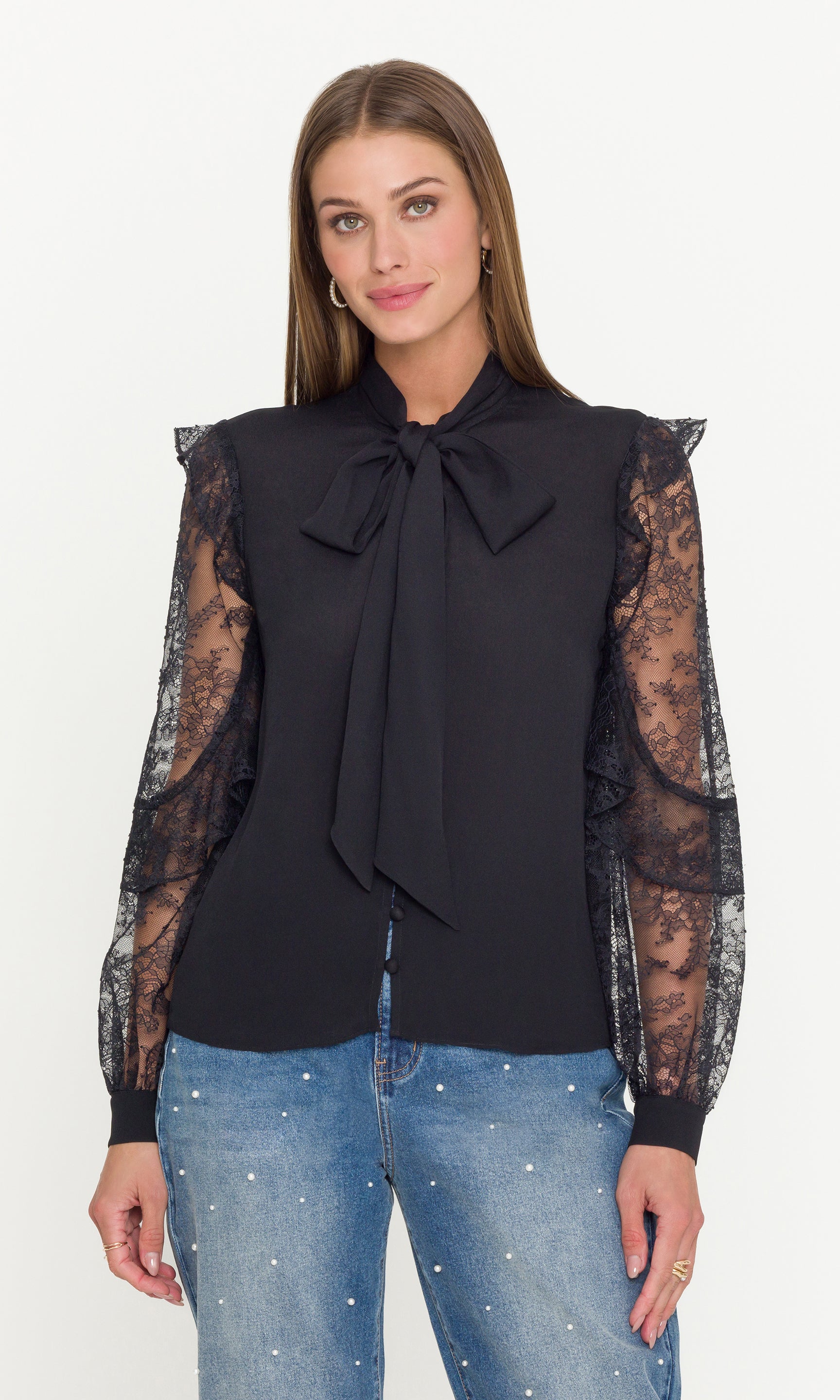 Woman wearing a black blouse with sheer sleeves and a bow tie, paired with blue jeans on a white background.