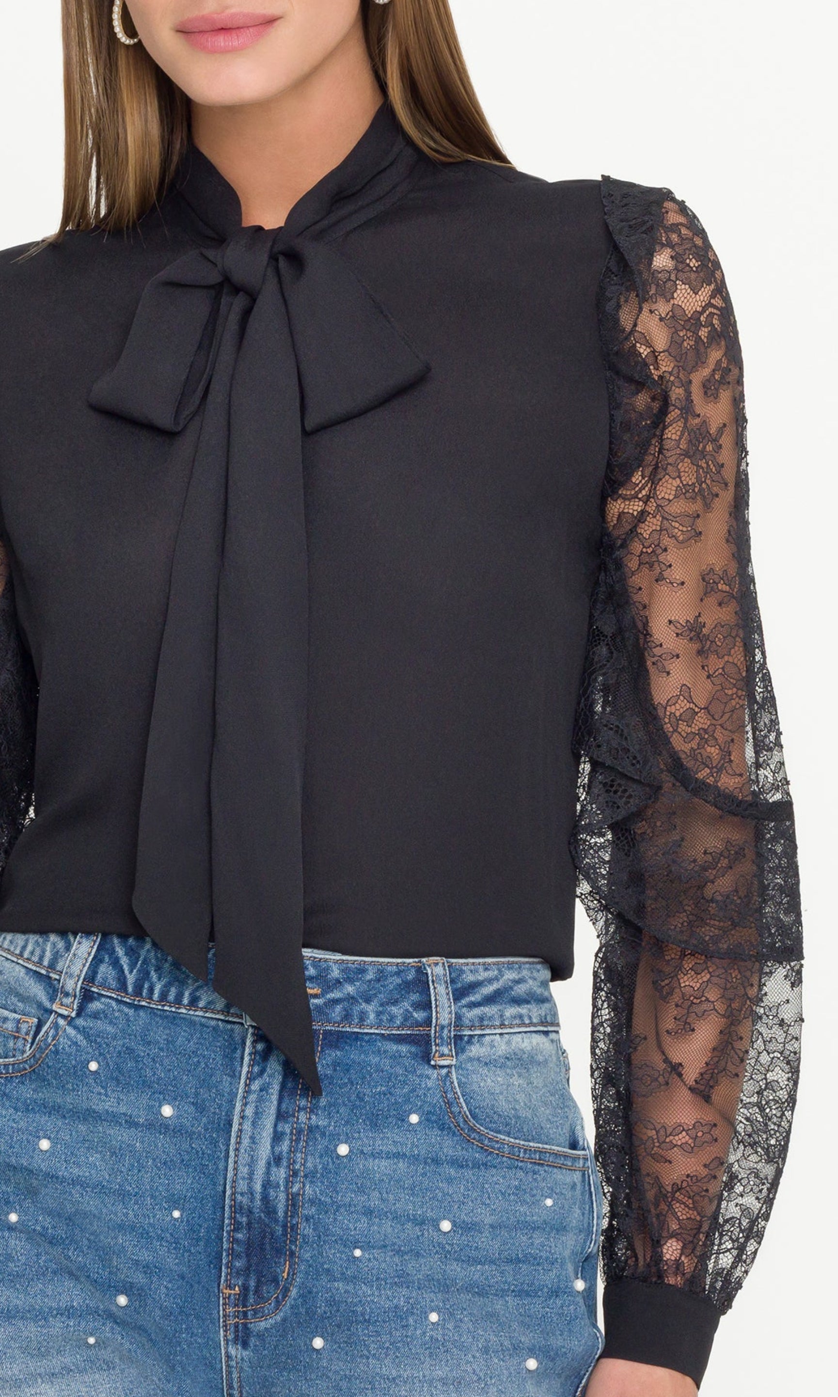 Woman wearing a black blouse with lace sleeves and a bow tie, paired with blue jeans on a white background.