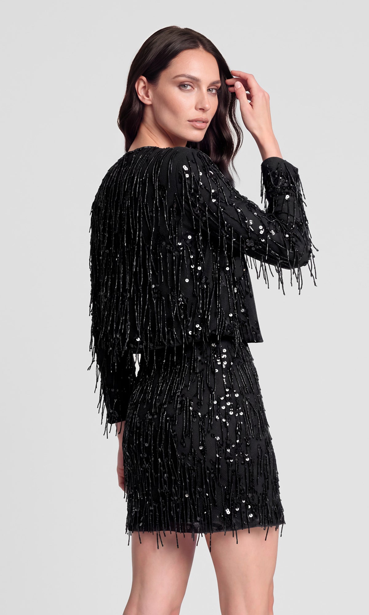 Back view of model wearing a black fringe sequins jacket over a matching fringe sequins skirt.