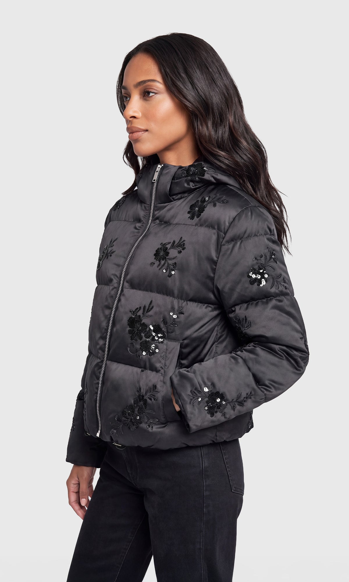 Side view of model wearing a black hooded puffer jacket with black floral embroidery and sequined details, paired with black pants.