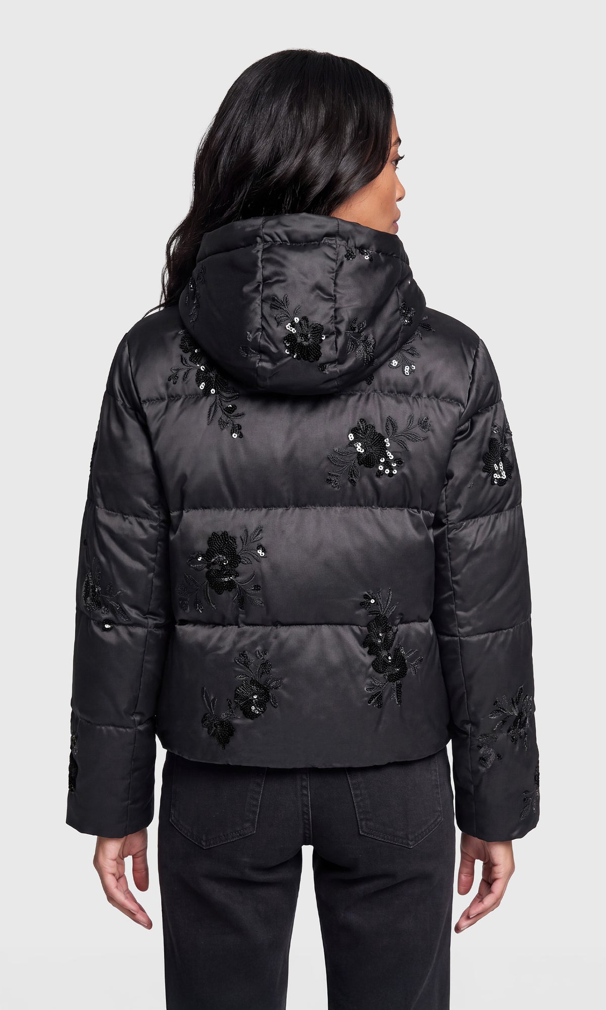 Back view of model wearing a black hooded puffer jacket with black floral embroidery and sequined details, paired with black pants.