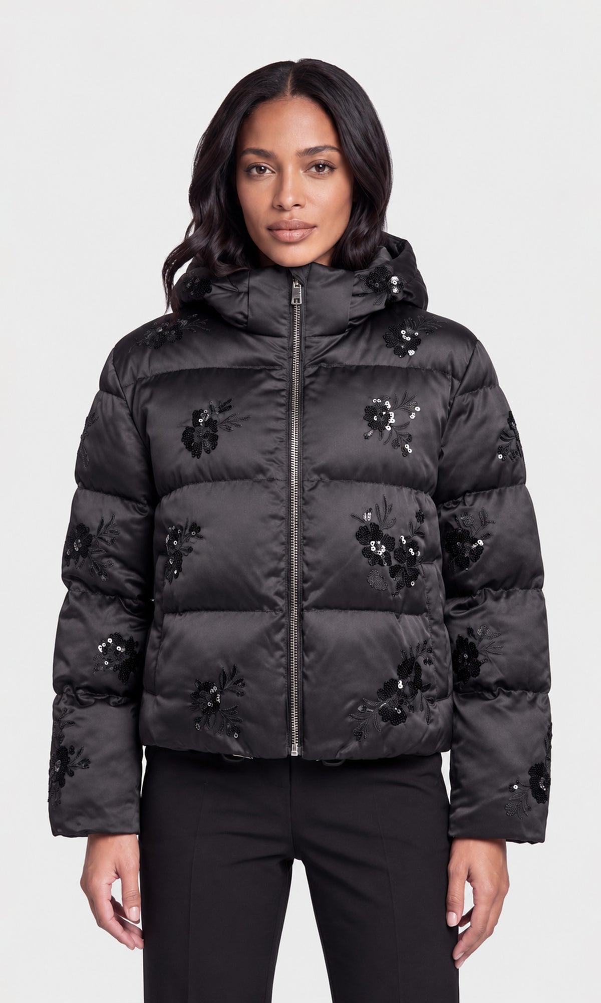 Model wearing a black hooded puffer jacket with black floral embroidery and sequined details, paired with black pants.